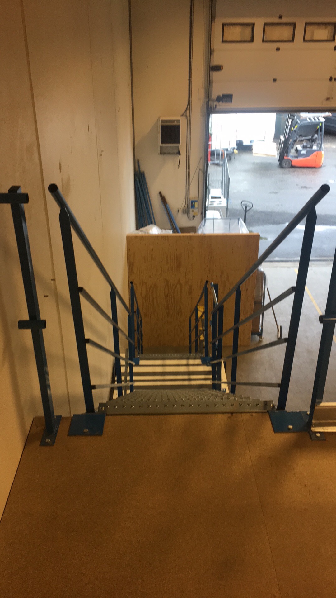Metalowe schody z niebieskimi barierkami prowadzące z antresoli w dół do hali magazynowej z widocznym wózkiem widłowym