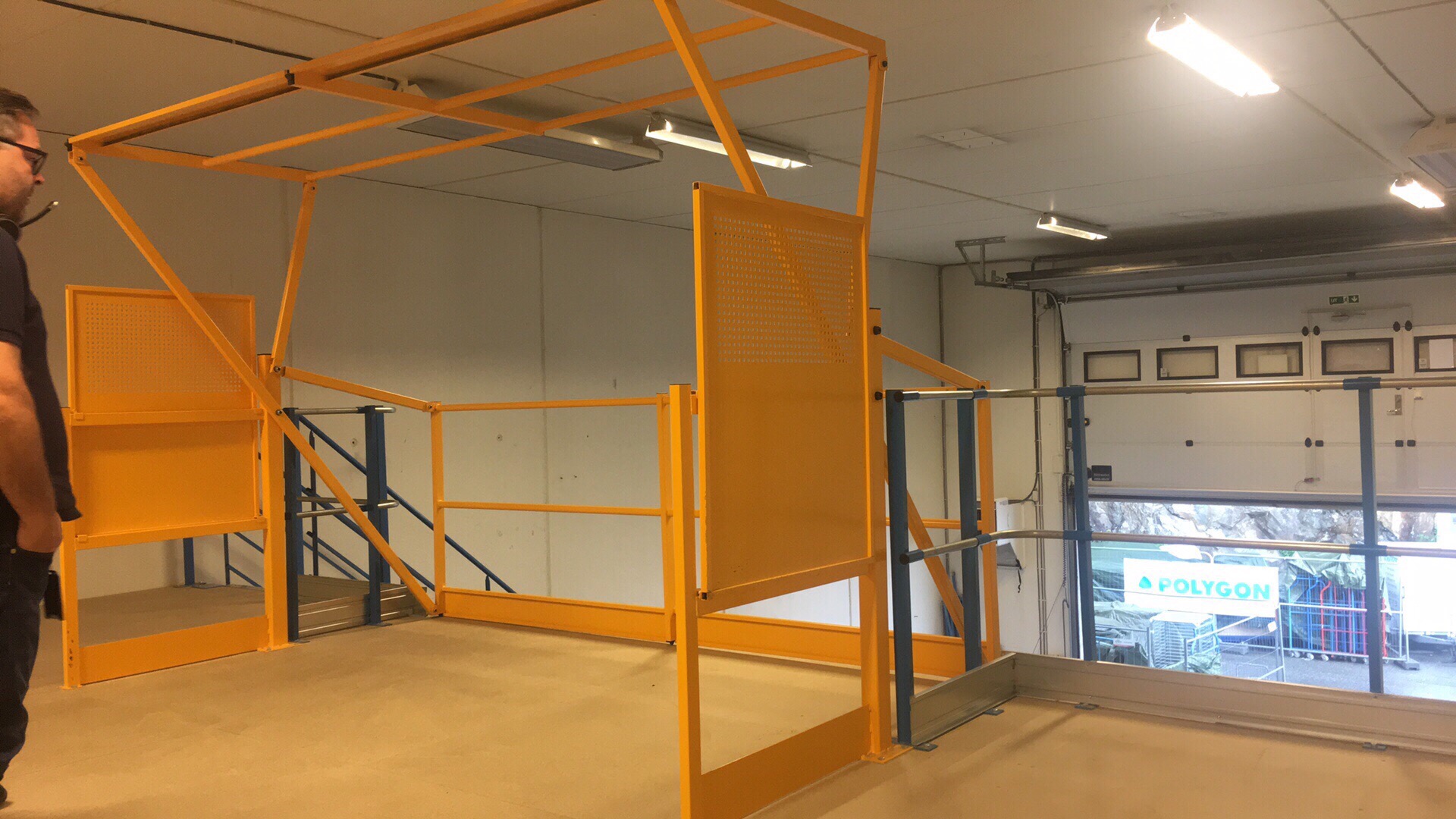 Żółta konstrukcja antresoli z perforowanymi panelami ochronnymi w magazynie, widoczny fragment sylwetki mężczyzny po lewej stronie.
