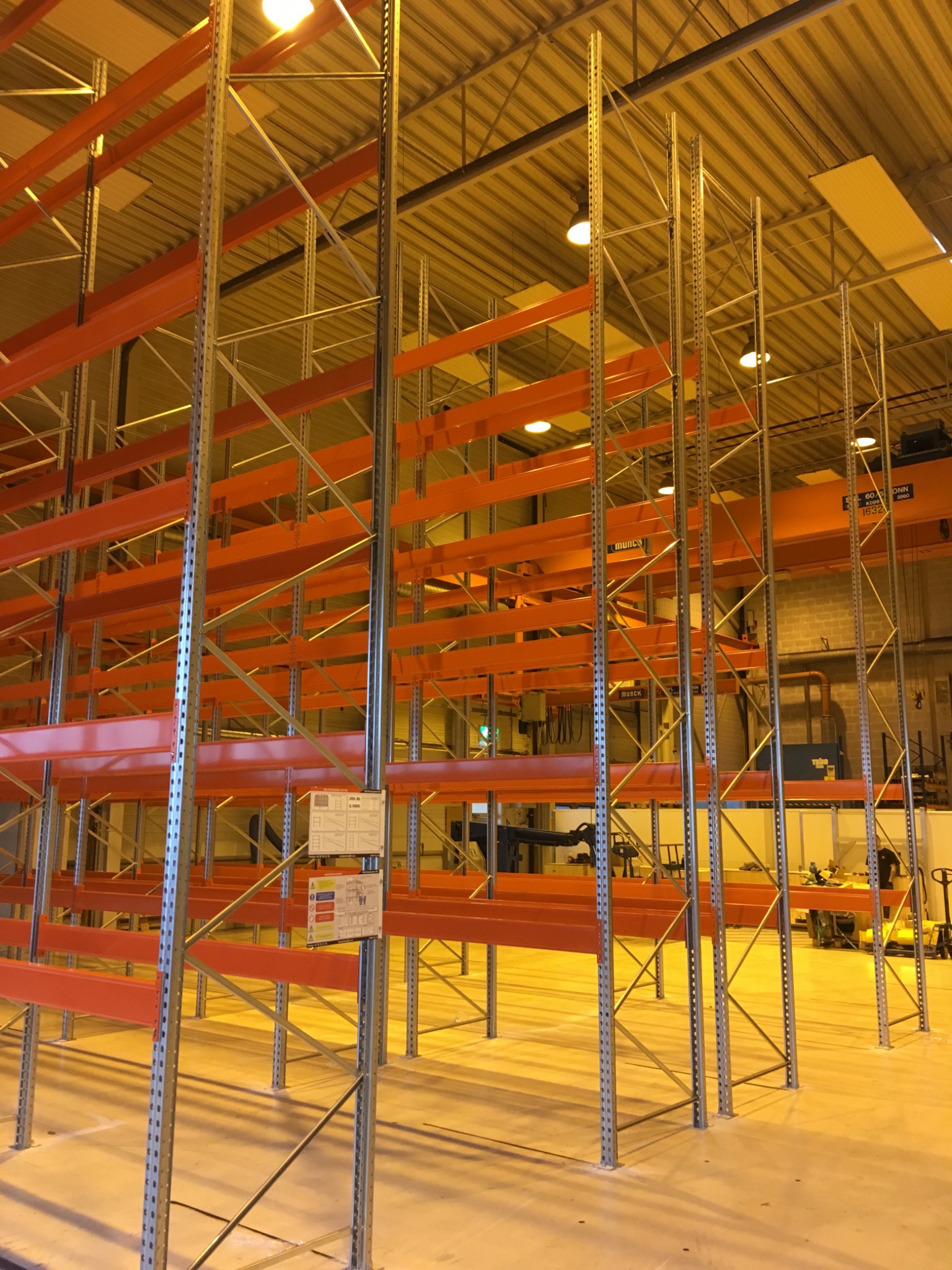 Metalowa konstrukcja regałów magazynowych z pomarańczowymi poprzeczkami w hali przemysłowej, widoczne tabliczki informacyjne na jednym z regałów.