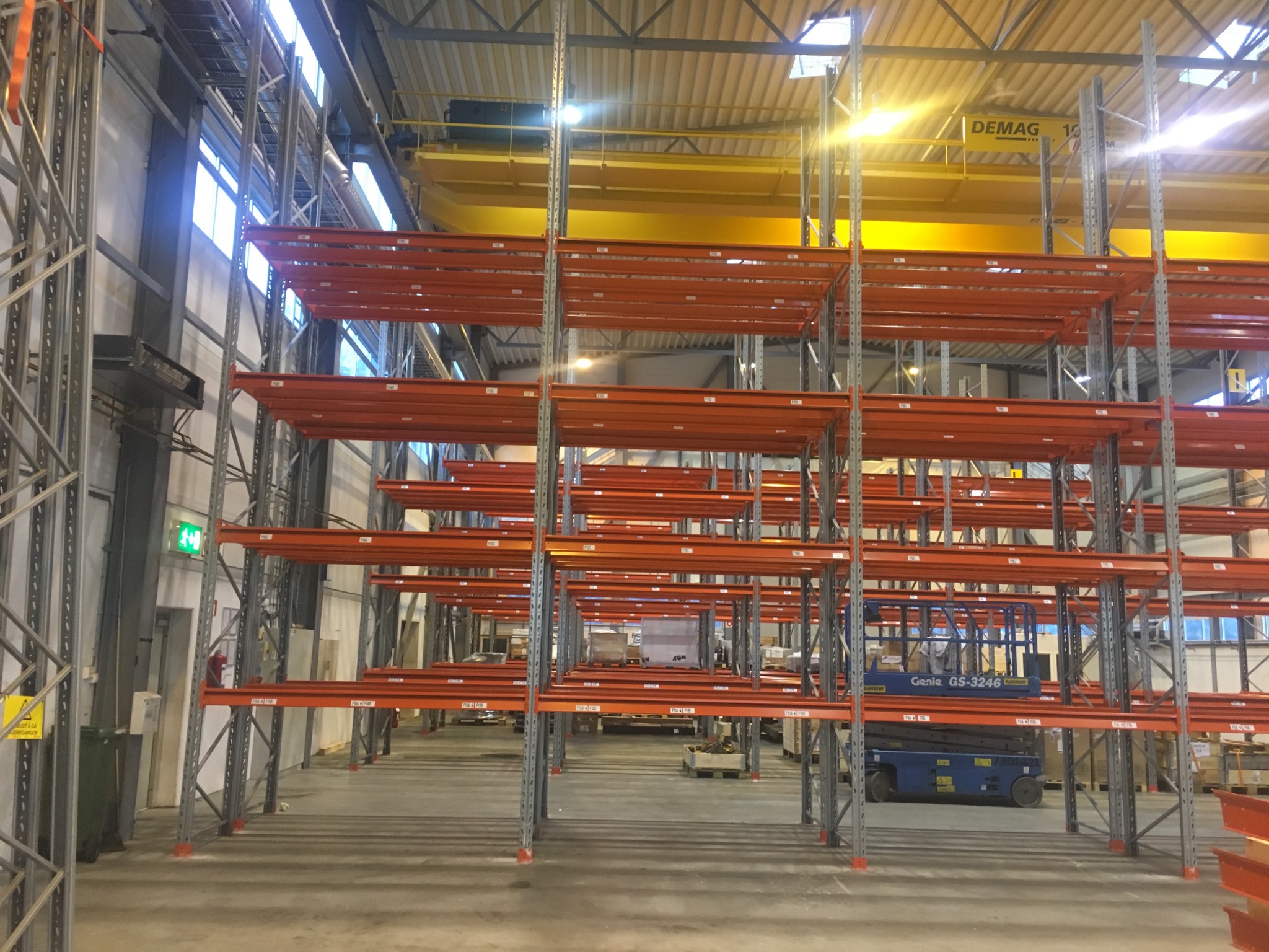 Wysokie, pomarańczowe metalowe regały w magazynie, widoczny podnośnik koszowy Genie GS-3246 i suwnica Demag pod sufitem.