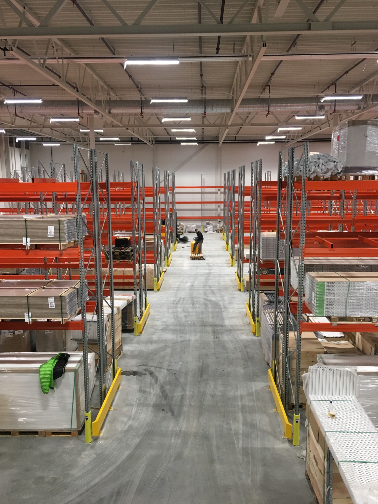 Wysokie regały magazynowe z pomarańczowymi belkami, wypełnione towarem, widok z góry na korytarz z pracownikiem na wózku widłowym w oddali.