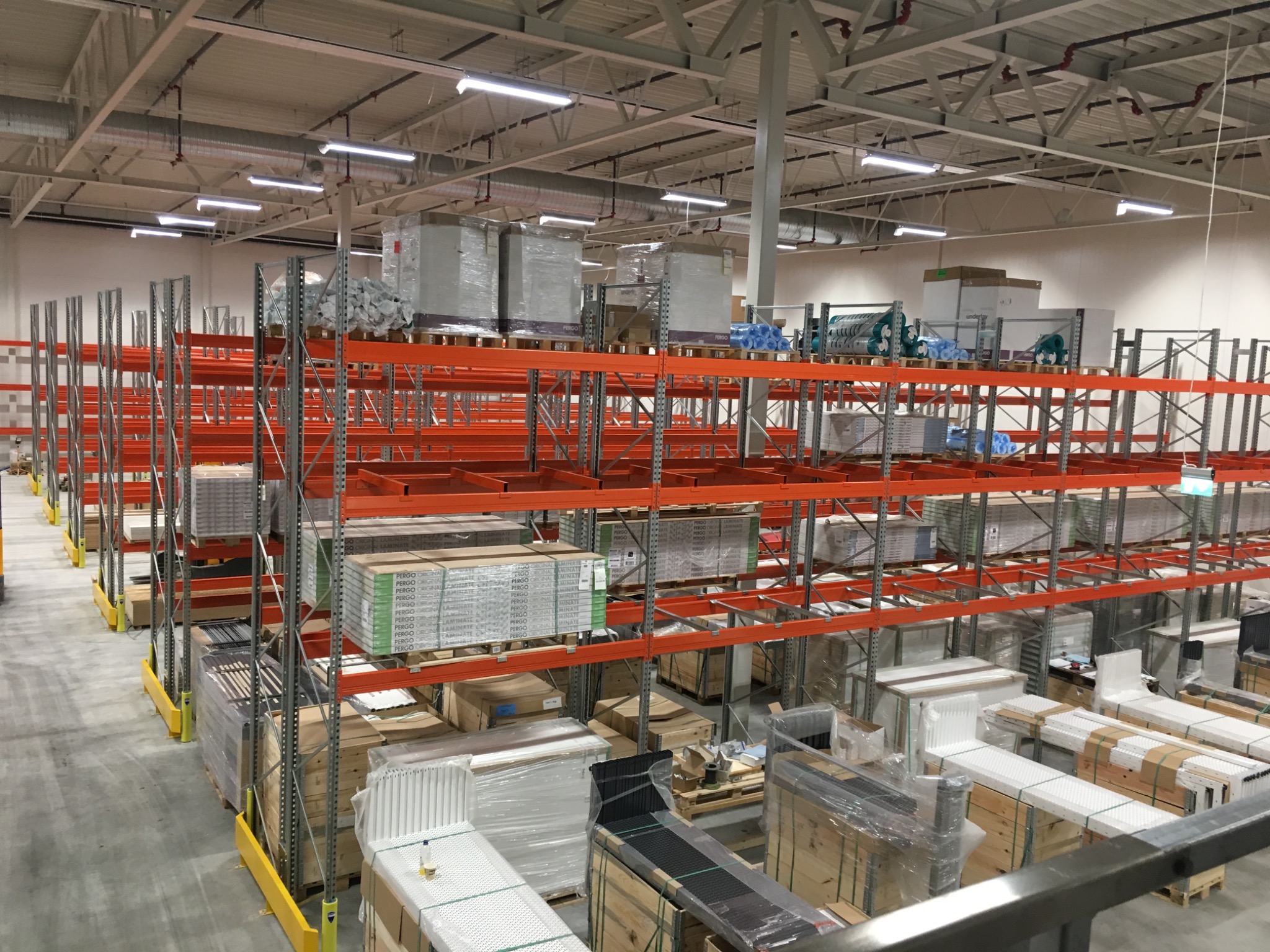 Wnętrze magazynu z pomarańczowymi regałami wypełnionymi różnorodnymi towarami, w tym paletami z panelami podłogowymi Pergo, zapakowanymi elementami i zabezpieczonymi folią stretch.