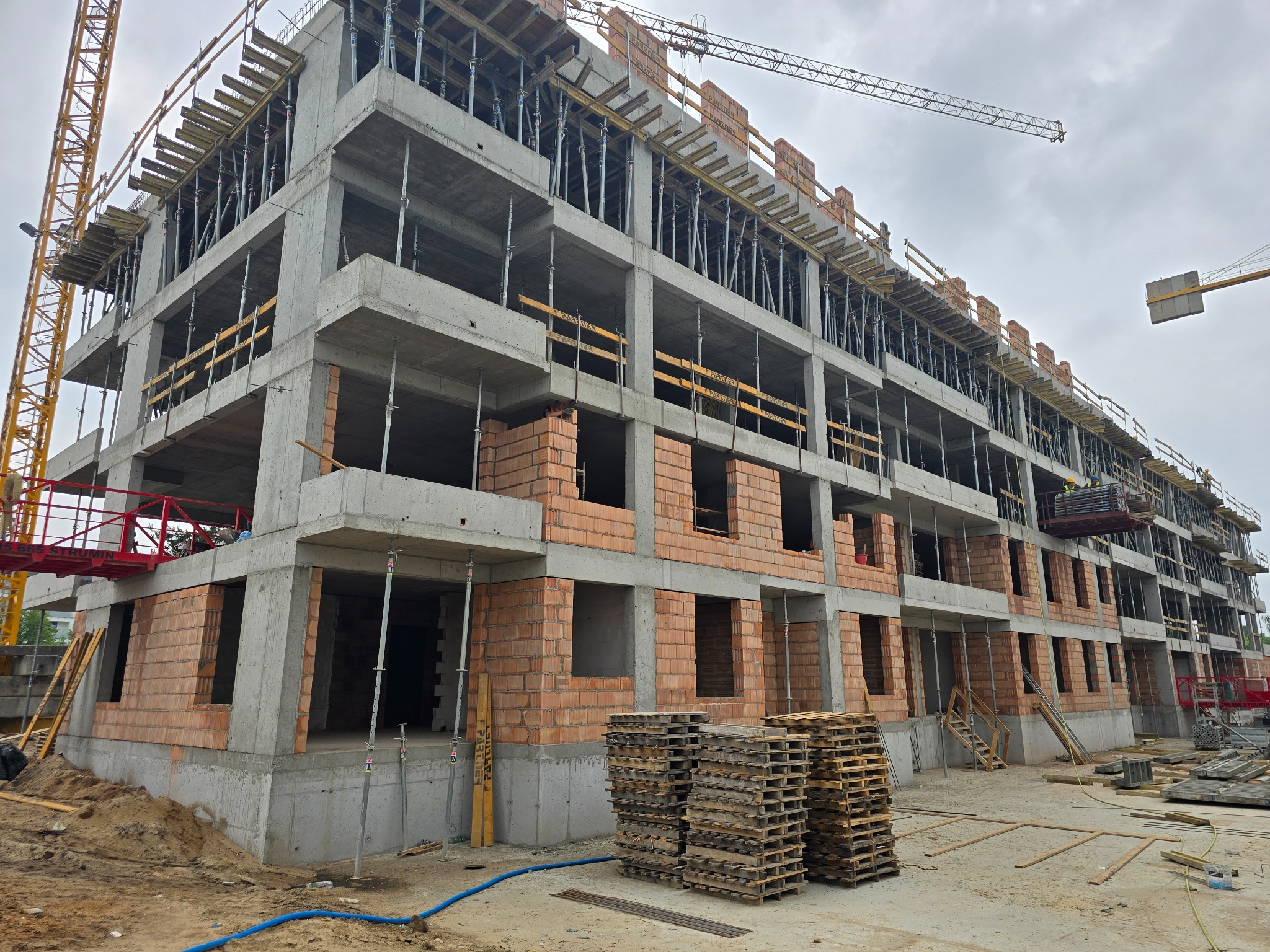 Wielopiętrowy budynek w stanie surowym otwartym, z widocznymi fragmentami ścian z cegieł, betonowymi słupami, drewnianymi szalunkami i dwiema wieżami dźwigów na pochmurnym niebie, z paletami...