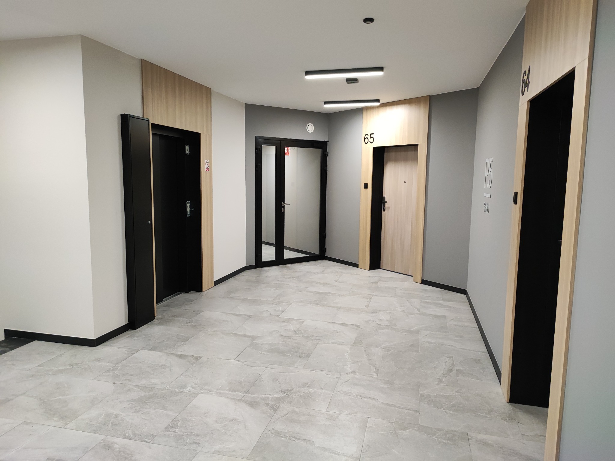 Nowoczesny korytarz w budynku mieszkalnym z szarymi ścianami, marmurową podłogą, drzwiami do mieszkań o numerach 64 i 65 oraz czarnymi framugami drzwi wejściowych.