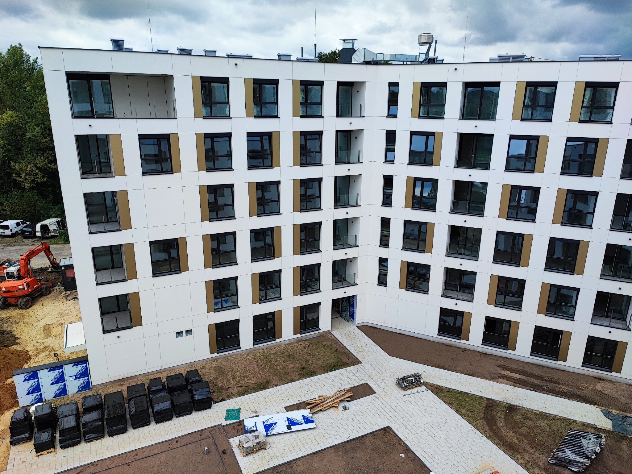 Wielopiętrowy budynek z białymi panelami i ciemnymi okiennicami w trakcie budowy, widoczny sprzęt budowlany i materiały na placu budowy, ułożona kostka brukowa na dziedzińcu.