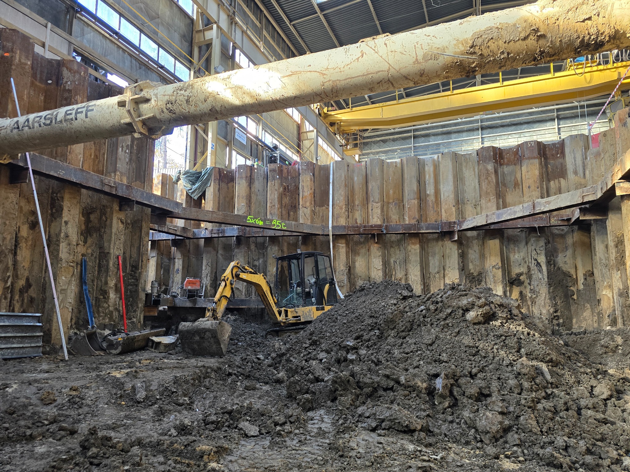 Żółta mini koparka w wykopie zabezpieczonym ściankami Larsena, widoczne narzędzia i rury konstrukcyjne pod stropem hali.