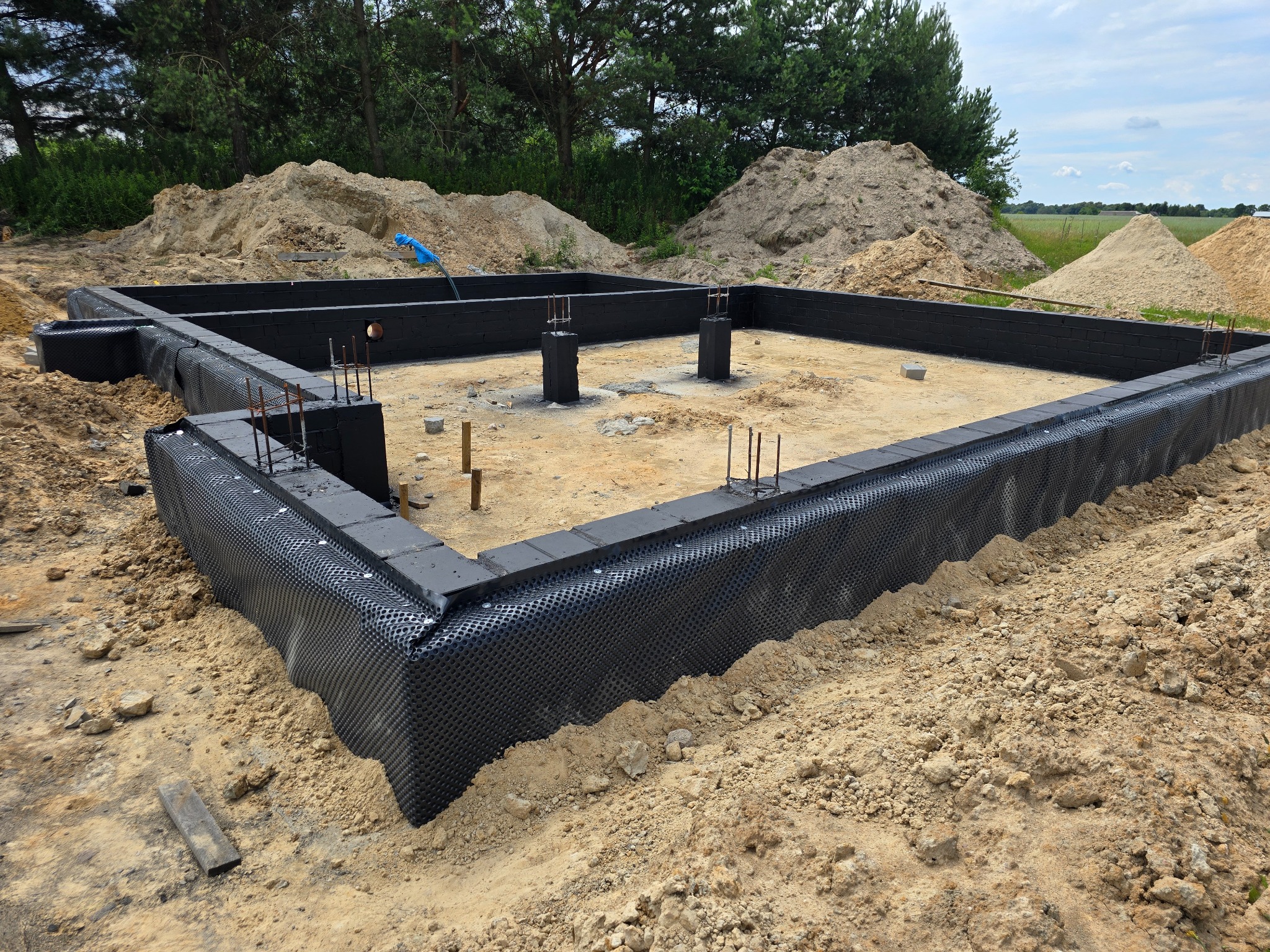 Fundamenty domu w trakcie budowy, zabezpieczone czarną folią hydroizolacyjną, z wystającymi prętami zbrojeniowymi, otoczone piaskiem i zalesionym terenem w tle.