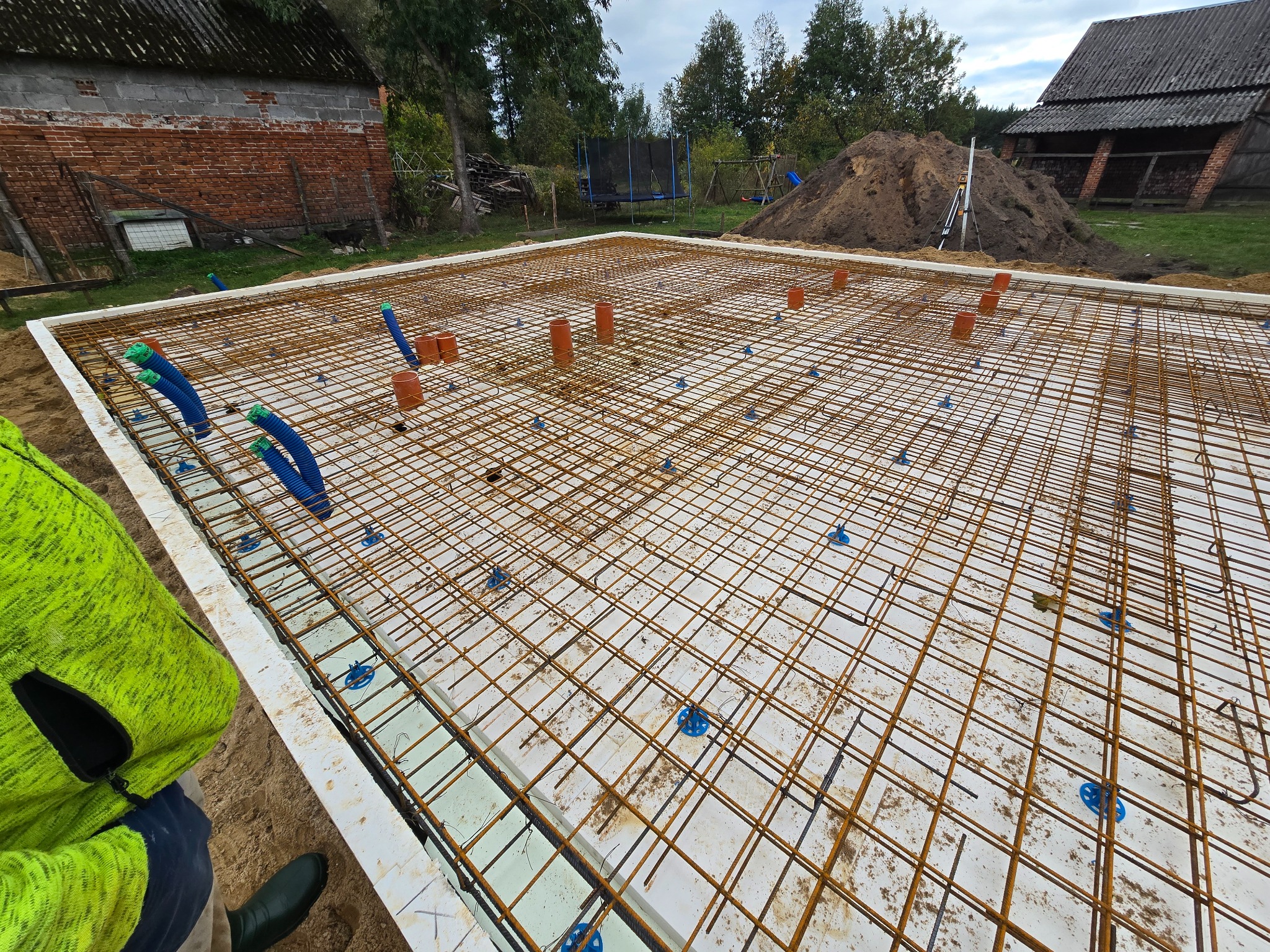 Zbrojenie fundamentów na białej izolacji, z wystającymi rurami kanalizacyjnymi, widok z góry na plac budowy w wiejskim otoczeniu.