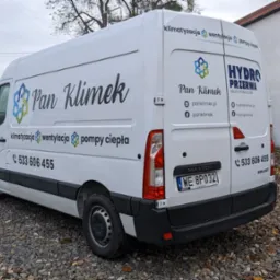 Biały samochód dostawczy z nadrukowanym logo firmy Pan Klimek oferującej usługi klimatyzacji, wentylacji i pomp ciepła, zaparkowany na żwirowym podłożu przed budynkiem.