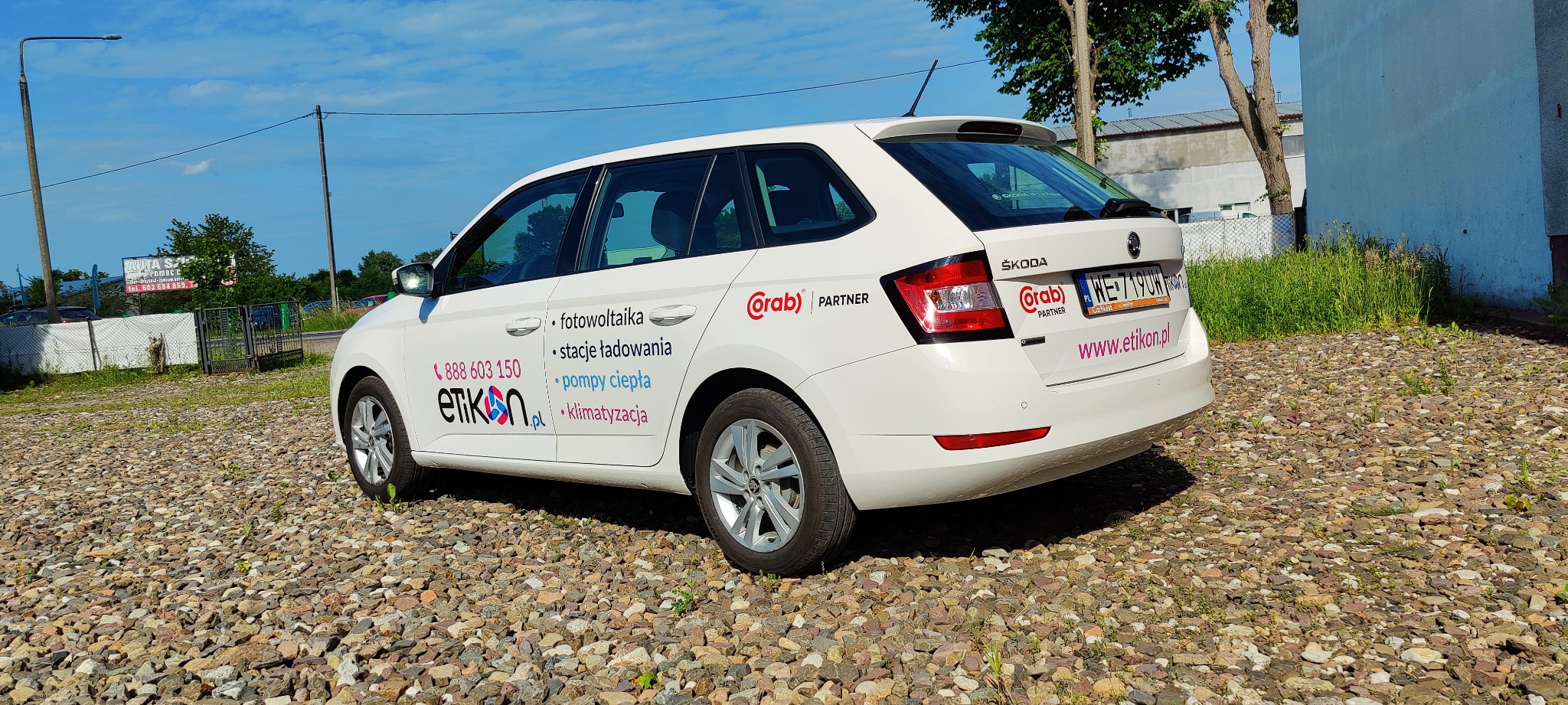 Biała Skoda Fabia z reklamą firmy Etikon na boku i z tyłu, oferującej fotowoltaikę, stacje ładowania, pompy ciepła i klimatyzację, zaparkowana na żwirowym terenie.