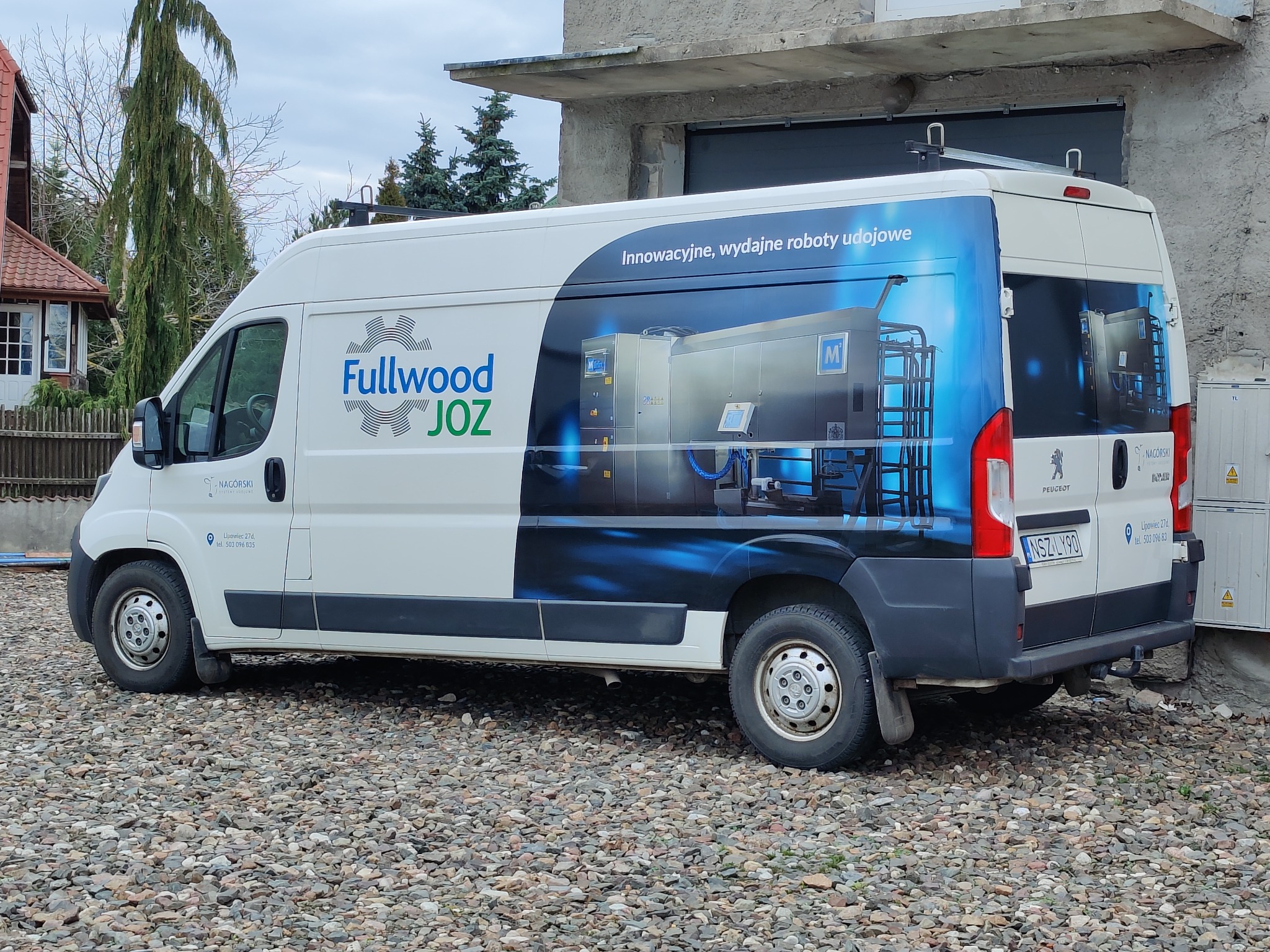 Biały samochód dostawczy z niebieskim nadrukiem reklamowym przedstawiającym automatyczne roboty udojowe oraz logo firmy Fullwood JOZ, zaparkowany na żwirowym podłożu przed budynkiem.