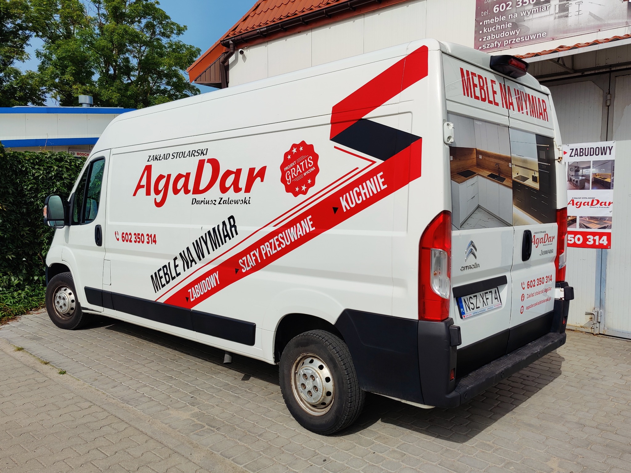 Biała furgonetka z reklamą zakładu stolarskiego AgaDar, prezentująca meble na wymiar, kuchnie i zabudowy, z numerem telefonu i adresem strony internetowej, zaparkowana na brukowanym placu.