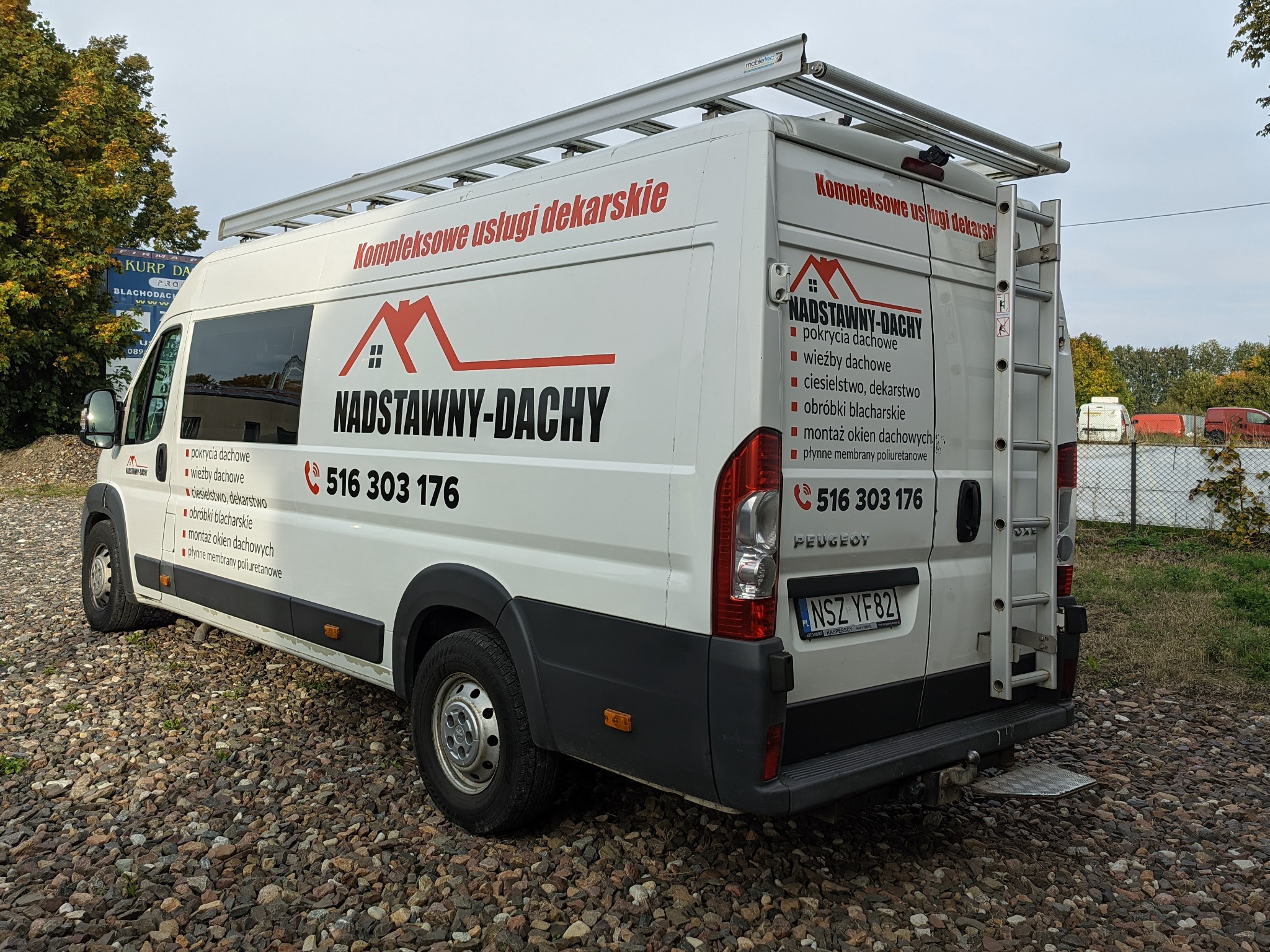Biały samochód dostawczy z logo firmy dekarskiej, widoczne dane kontaktowe i zakres usług, drabina na dachu i z tyłu pojazdu, zaparkowany na żwirowym podłożu.