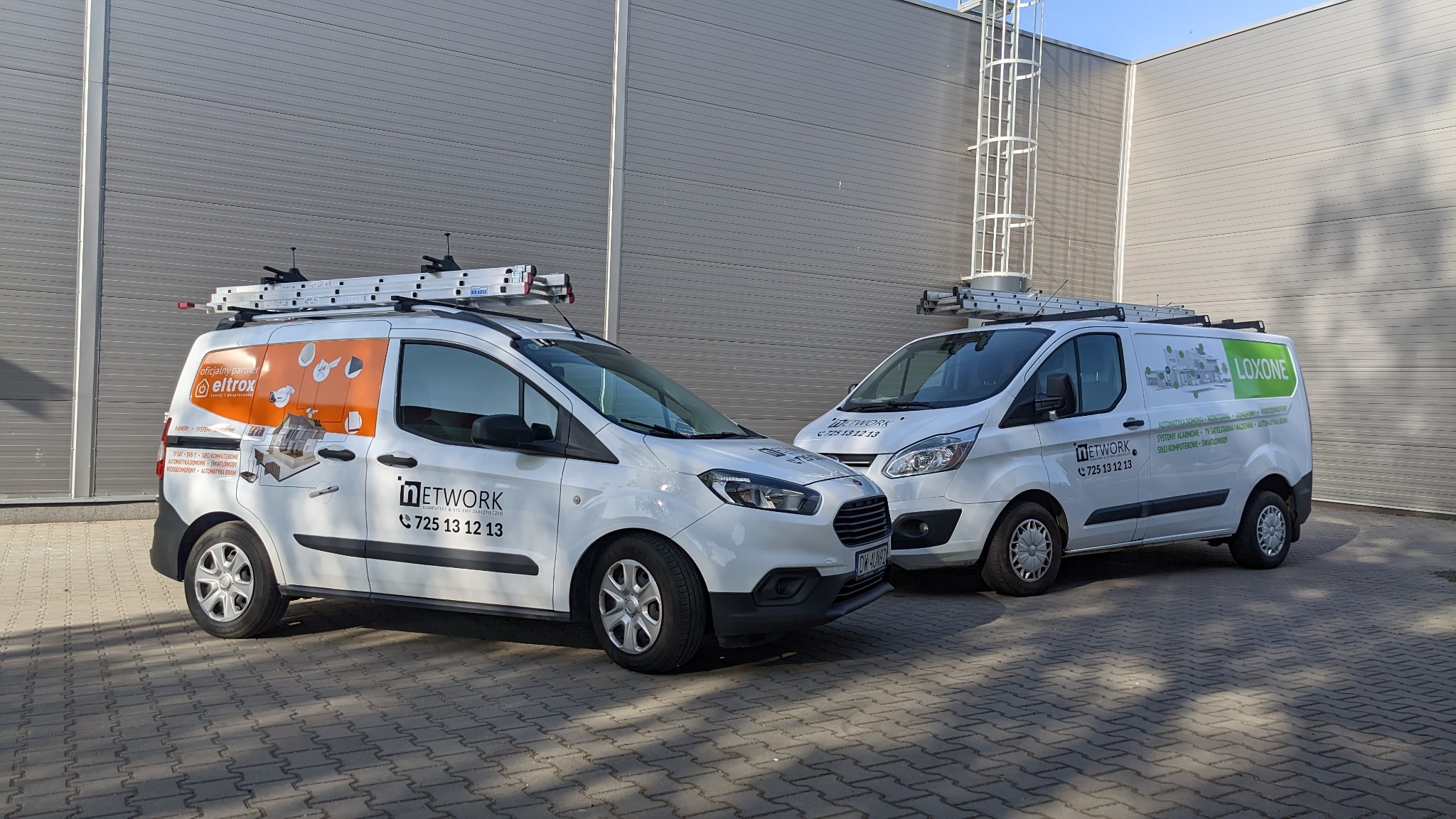 Dwa białe samochody dostawcze z logo firm instalacyjnych i drabinami na dachu, zaparkowane przed szarą ścianą budynku.