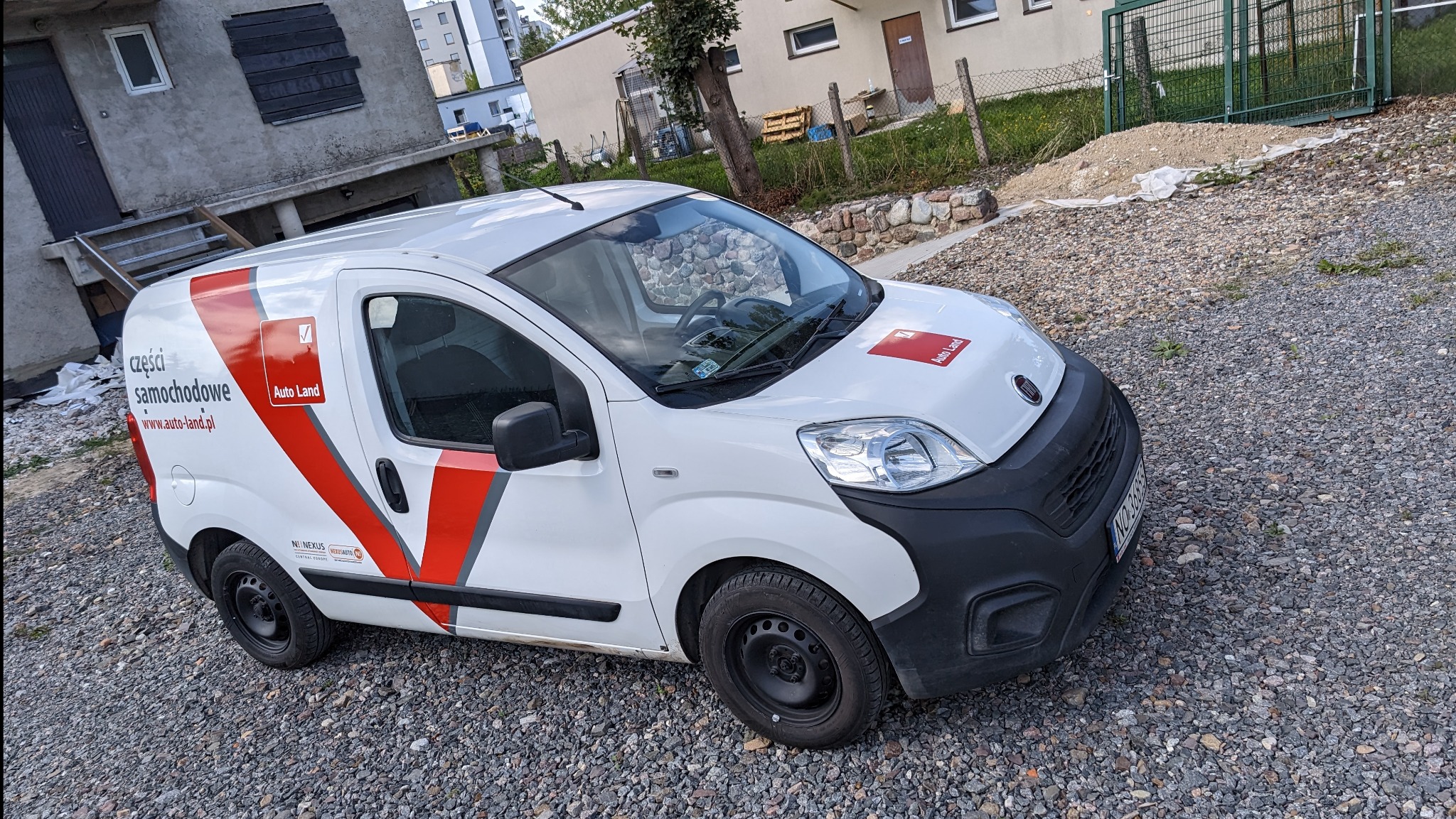 Biały samochód dostawczy z czerwonymi elementami graficznymi i logo firmy Auto Land, zaparkowany na żwirowym podłożu w otoczeniu budynków.