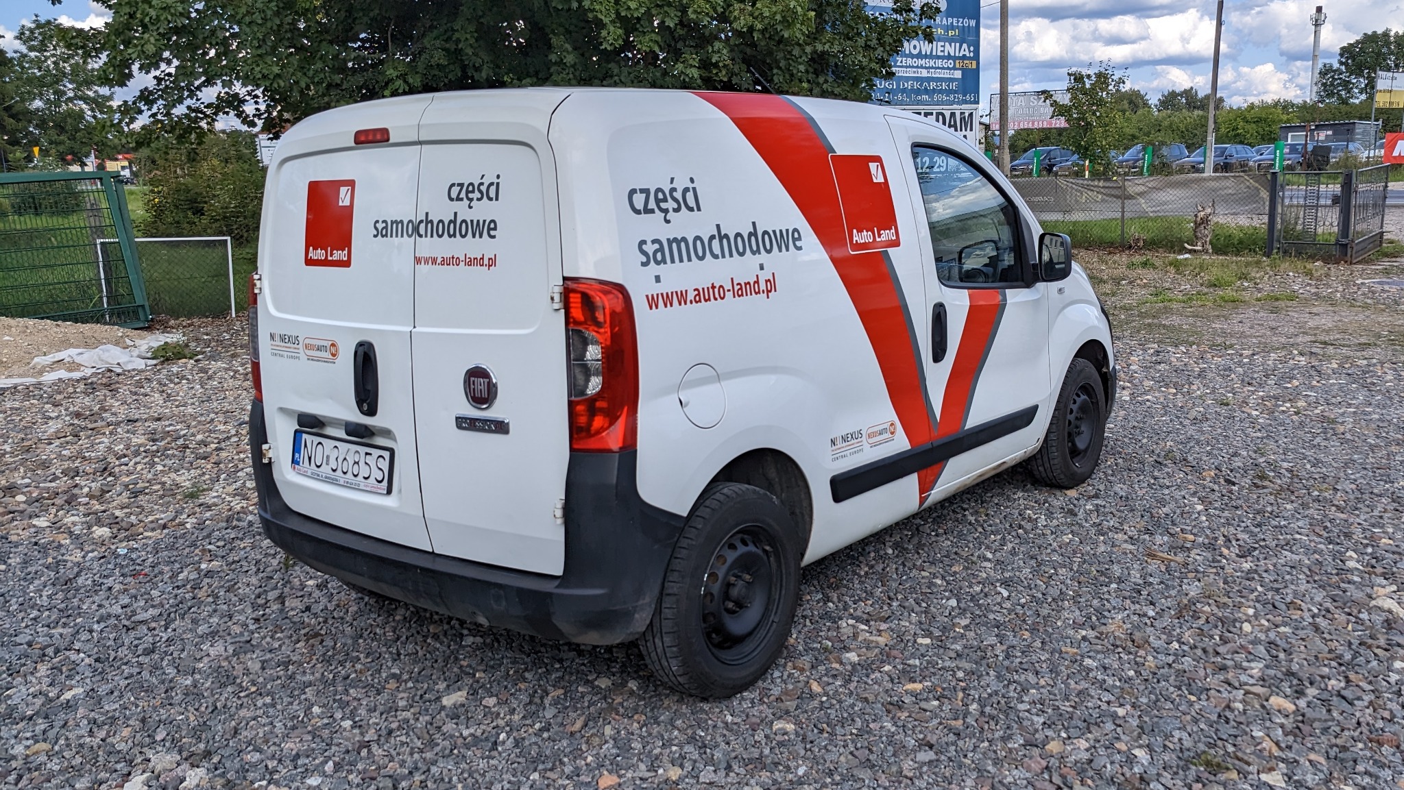 Biały samochód dostawczy z czerwonymi elementami graficznymi i logotypem firmy z branży motoryzacyjnej 'Auto Land' na parkingu w Szczytnie.