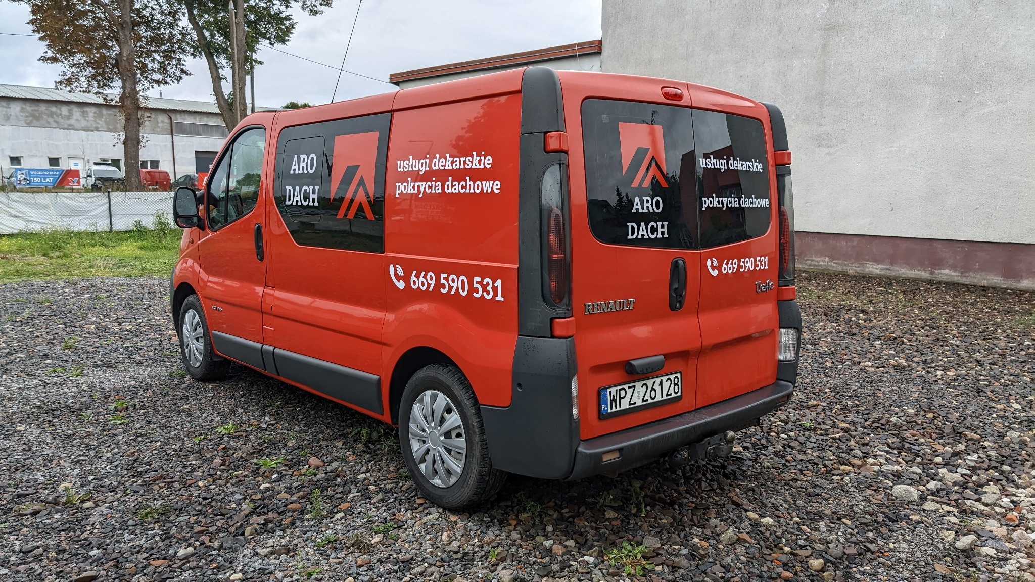 Pomarańczowy samochód dostawczy Renault z czarnymi napisami 'ARO DACH usługi dekarskie pokrycia dachowe' oraz numerem telefonu, zaparkowany na żwirowym podłożu.