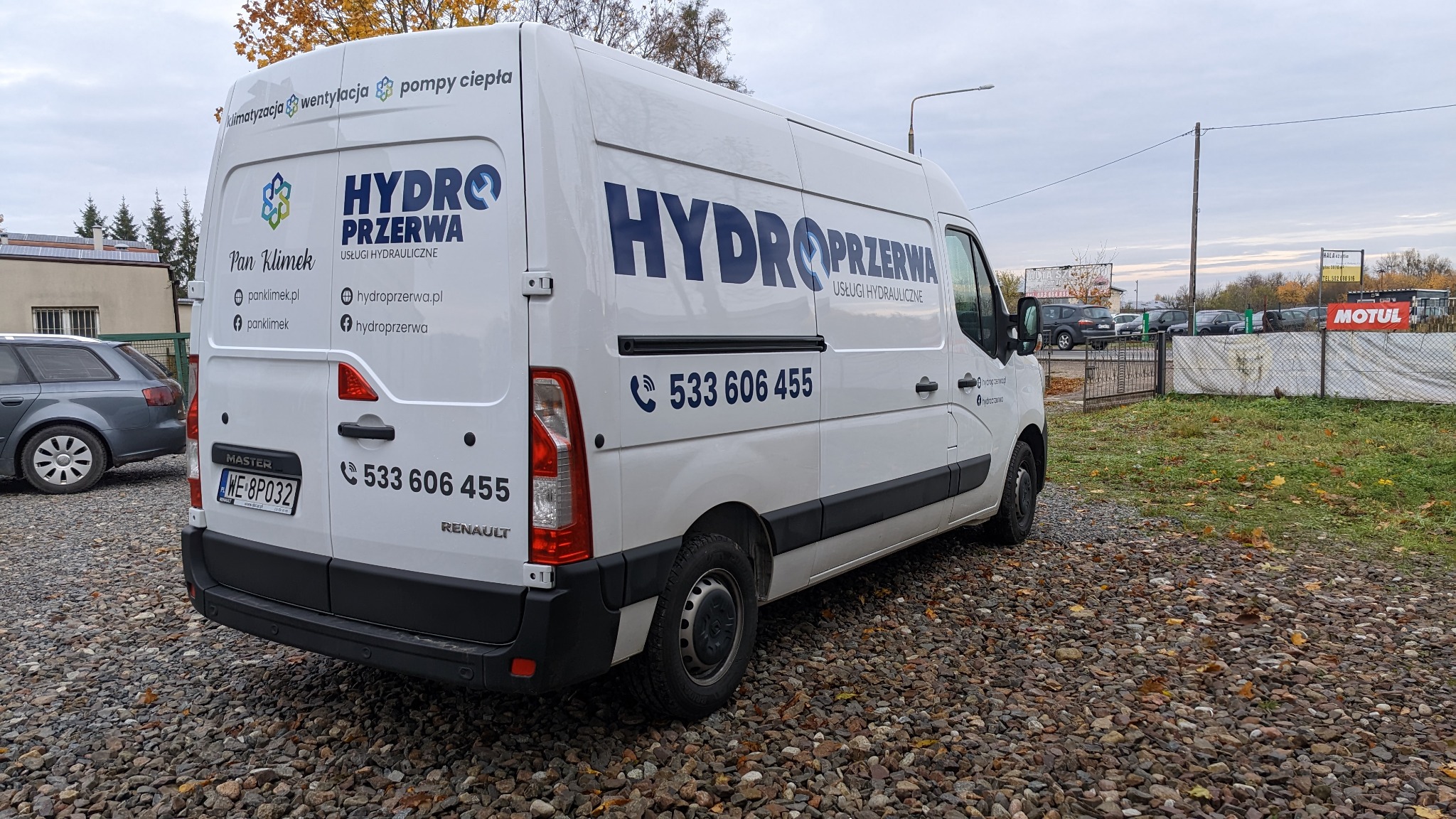 Biały samochód dostawczy Renault Master z reklamą usług hydraulicznych Hydro Przerwa, zaparkowany na żwirowym parkingu w pochmurny dzień.