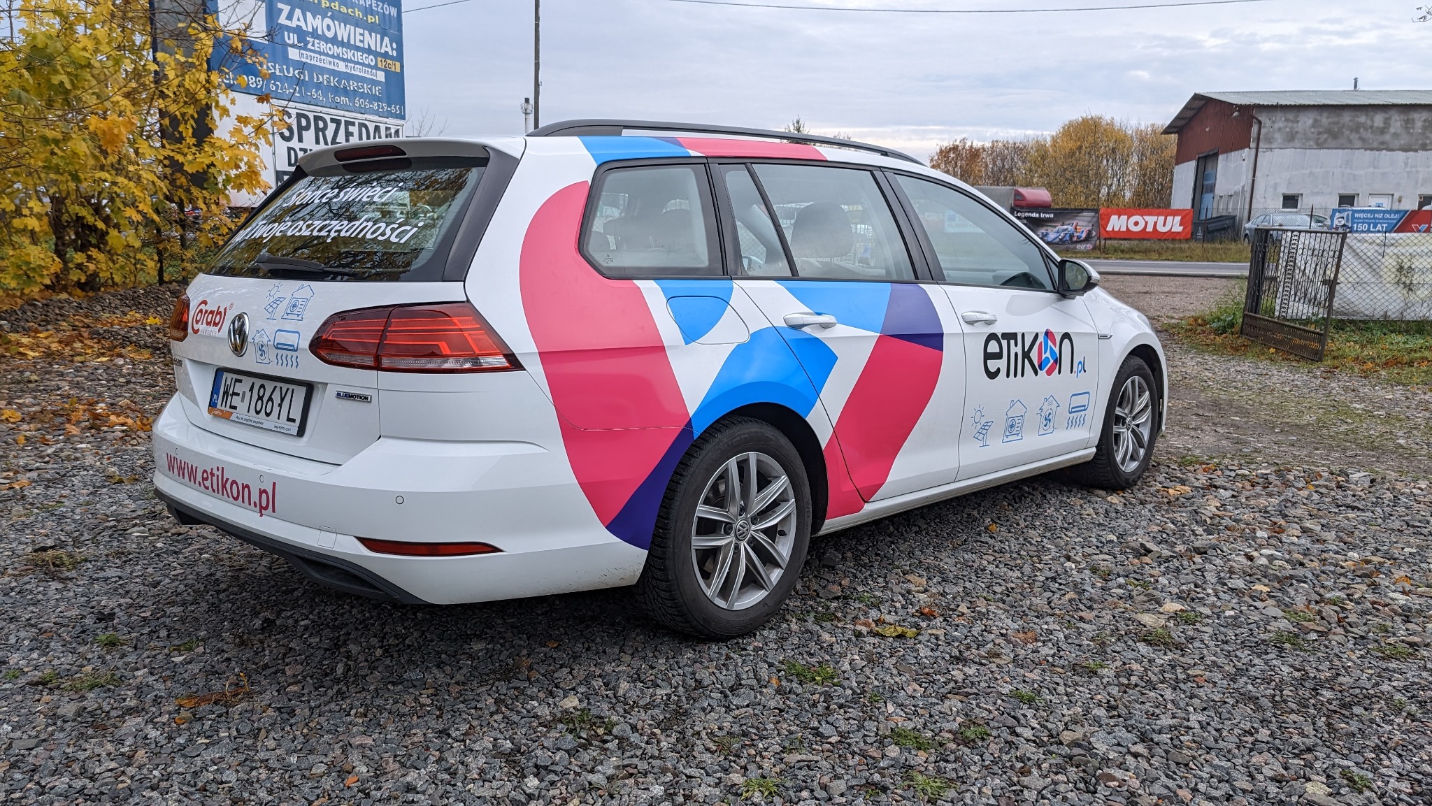 Biały samochód kombi z kolorowymi, geometrycznymi nadrukami w odcieniach różu i niebieskiego, zaparkowany na żwirowym parkingu. Na boku widoczne logo i ikony związane z branżą ekologiczną.