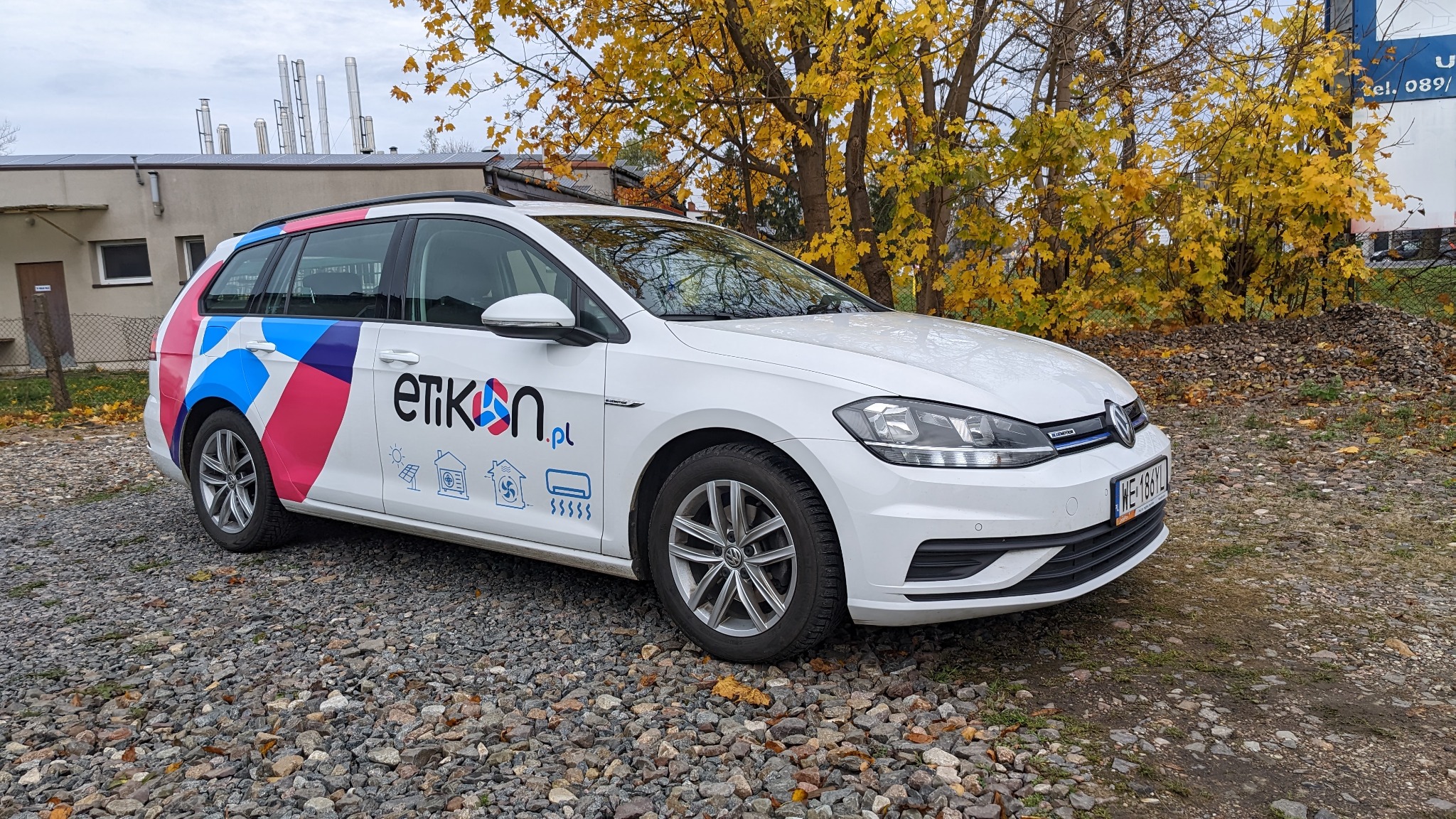 Biały samochód z oklejonym logo firmy 'etikon.pl' i grafikami instalacji solarnych, klimatyzacji i ogrzewania, zaparkowany na żwirowym podłożu z jesiennymi liśćmi.