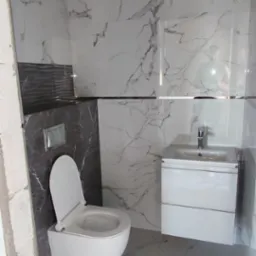 Nowoczesna łazienka w trakcie wykańczania, z białą wiszącą toaletą, umywalką z szafką i marmurowymi płytkami na ścianach i podłodze, widoczne niedokończone obramowanie drzwi.