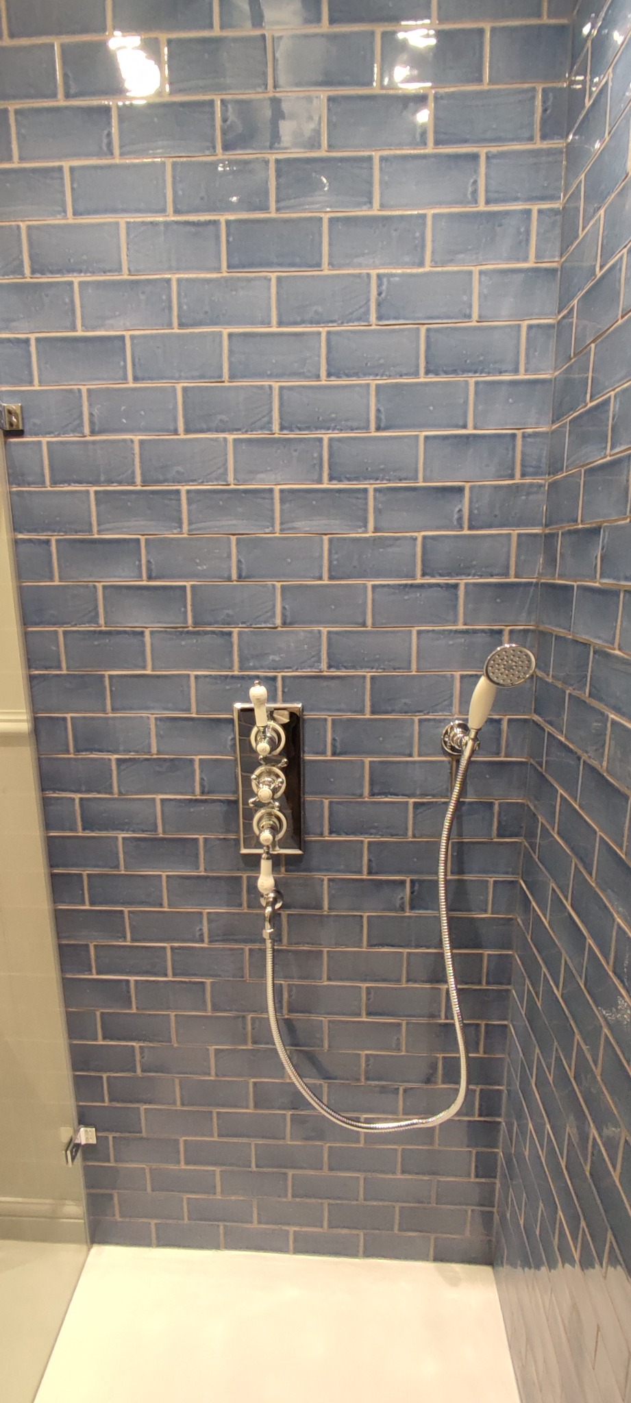 Niebieskie, błyszczące kafelki ułożone jak cegły w kabinie prysznicowej, z chromowaną baterią i słuchawką prysznicową w stylu retro.