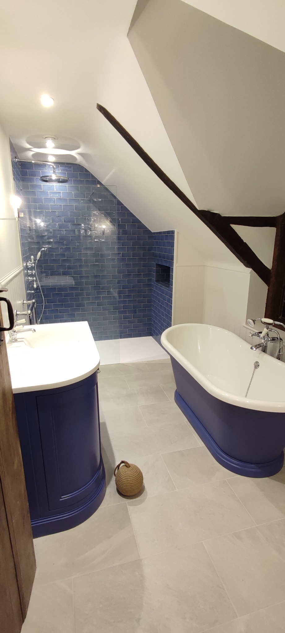 Wykończona łazienka na poddaszu ze skośnym sufitem, z kabiną prysznicową z niebieskimi płytkami, wolnostojącą wanną w kolorze granatowym, umywalką z szafką w tym samym odcieniu, szarą podłogą...