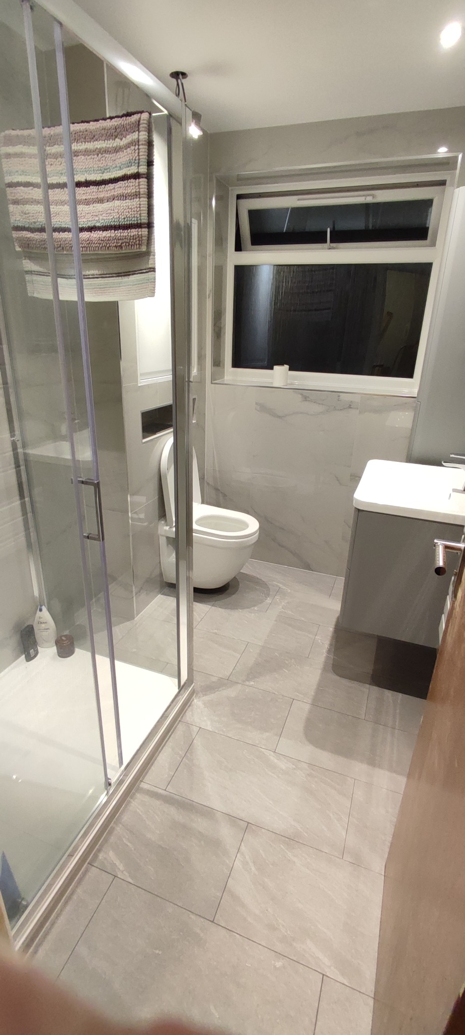 Jasna łazienka z kabiną prysznicową, toaletą i umywalką w szafce, wykończona płytkami imitującymi marmur, z ręcznikiem na kabinie.