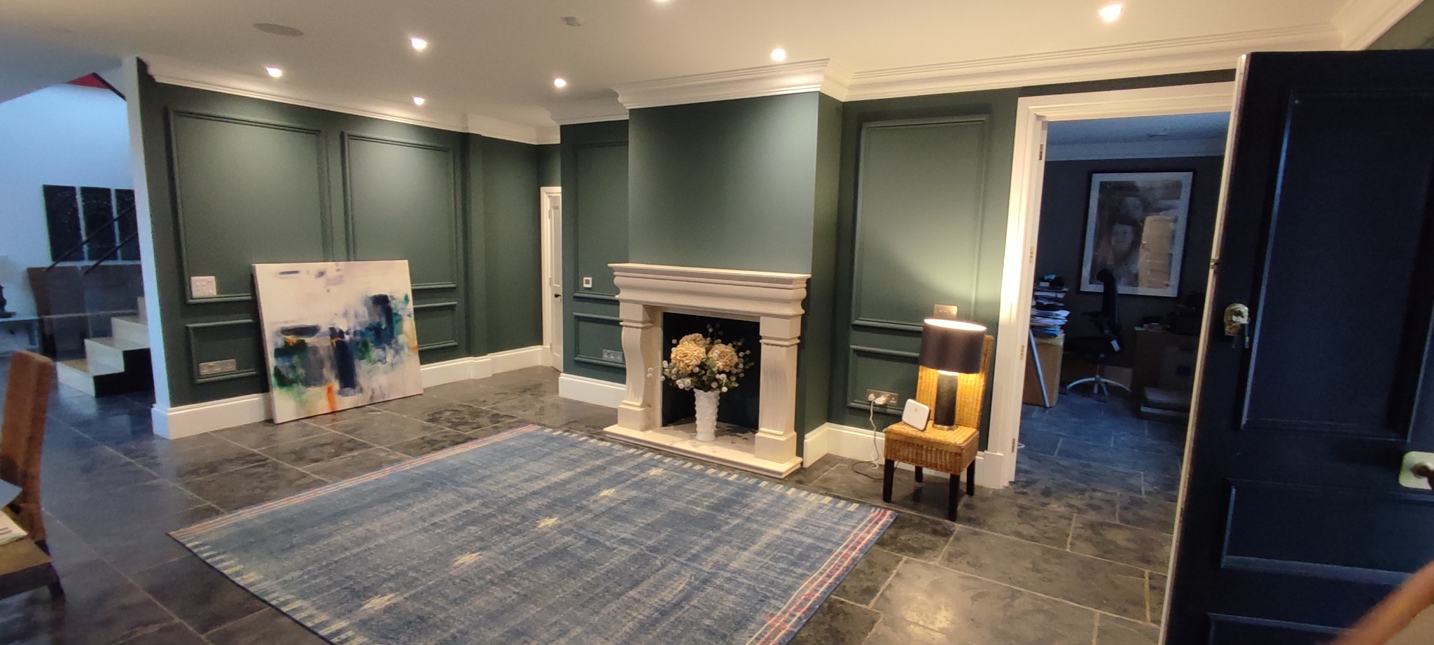 Eleganckie wnętrze z ciemnozielonymi ścianami, sztukaterią, kamiennym kominkiem z kwiatami, abstrakcyjnym obrazem i niebieskim dywanem w stylu vintage.