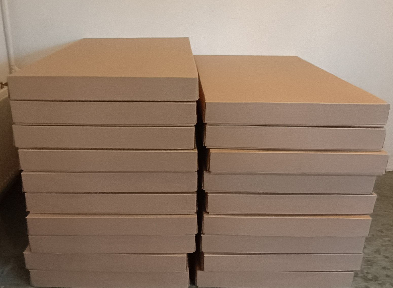 Dwa stosy płaskich, tekturowych pudeł w kolorze naturalnego brązu, ułożone jeden obok drugiego na szarej posadzce, widoczne na tle białej ściany.