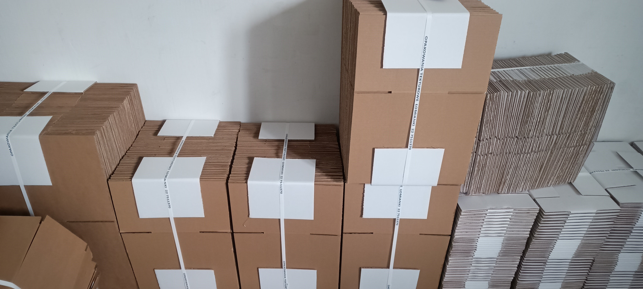 Stosy brązowych kartonowych pudeł, niektóre związane białą taśmą z nadrukowanym tekstem, ułożone na sobie i obok siebie pod białym sufitem, widać krawędzie tektury falistej.