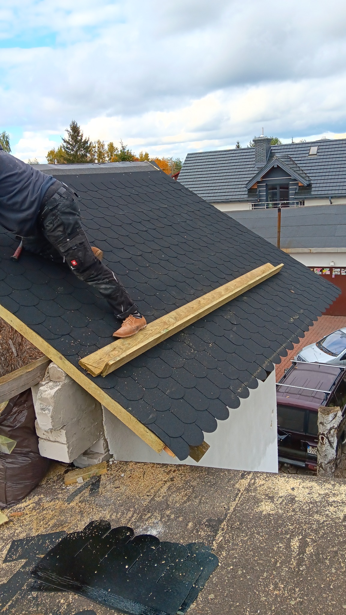 Dekarz w spodniach roboczych marki Engelbert Strauss i brązowych butach na drewnianej łacie montuje gont bitumiczny na dachu, widoczne samochody w tle.