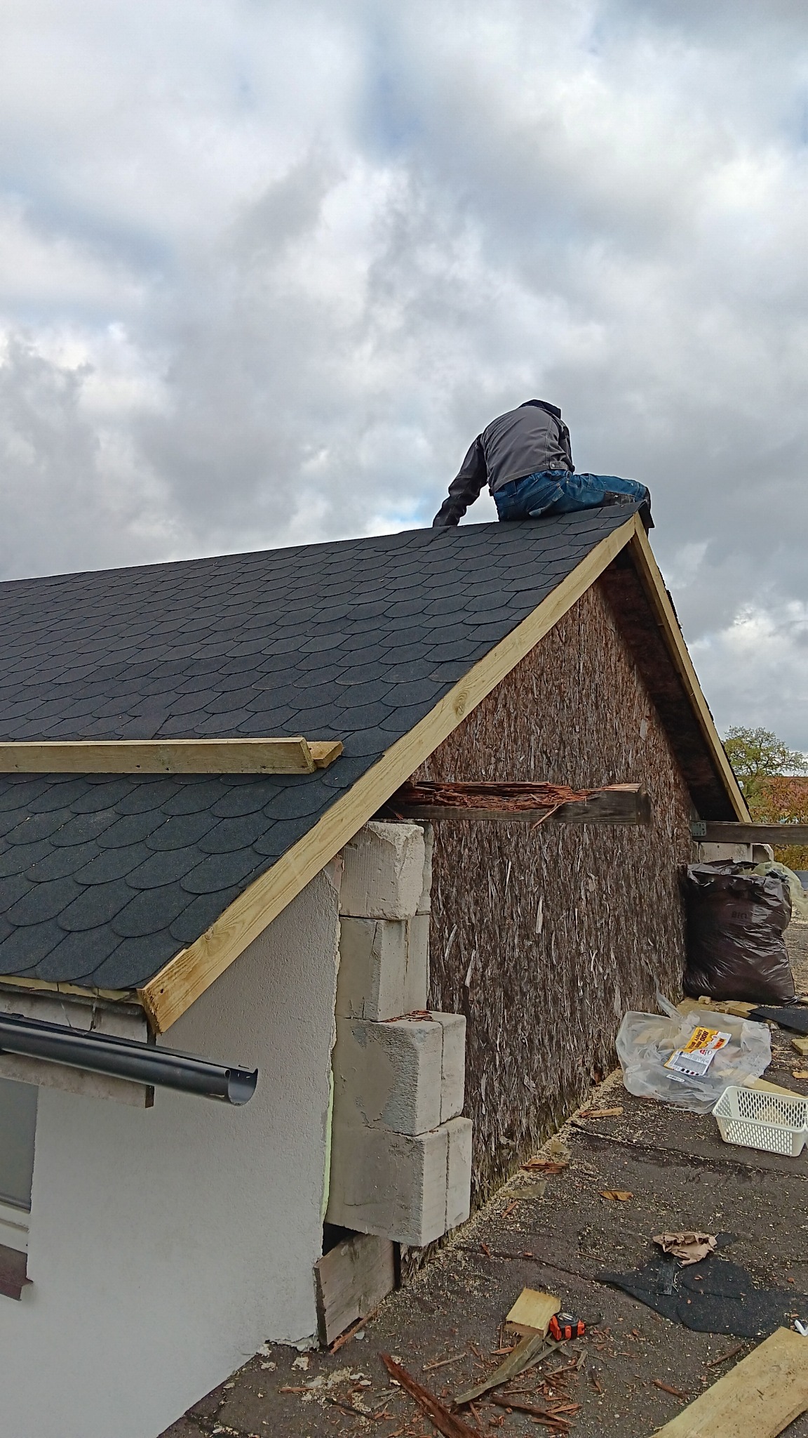 Pracownik siedzi na szczycie dachu pokrytego czarnym gontem bitumicznym w trakcie prac w Warszawie, widoczne elementy konstrukcyjne dachu i rynna.