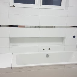 MK Renoma - Biała łazienka z wanną obudowaną płytkami, chromowane listwy dekoracyjne, okno w górnej części ściany, widoczne przyłącza hydrauliczne.