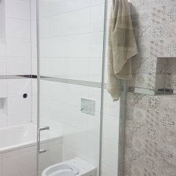 MK Renoma - Nowoczesna łazienka z kabiną prysznicową ze szkła hartowanego, wisząca toaleta, białe i wzorzyste płytki oraz beżowy ręcznik na relingu.