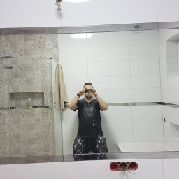 MK Renoma - Odbicie w lustrze łazienki po remoncie, widoczna kabina prysznicowa z wzorzystymi płytkami, białe płytki ścienne z metalowymi listwami, okno, oraz osoba robiąca zdjęcie telefonem, ubrana w robocze...