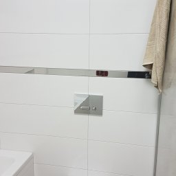 MK Renoma - Biała wisząca toaleta bez deski, zamontowana na ścianie z białych płytek z chromowaną listwą dekoracyjną i przyciskiem spłukującym, obok fragment wanny i beżowy ręcznik.