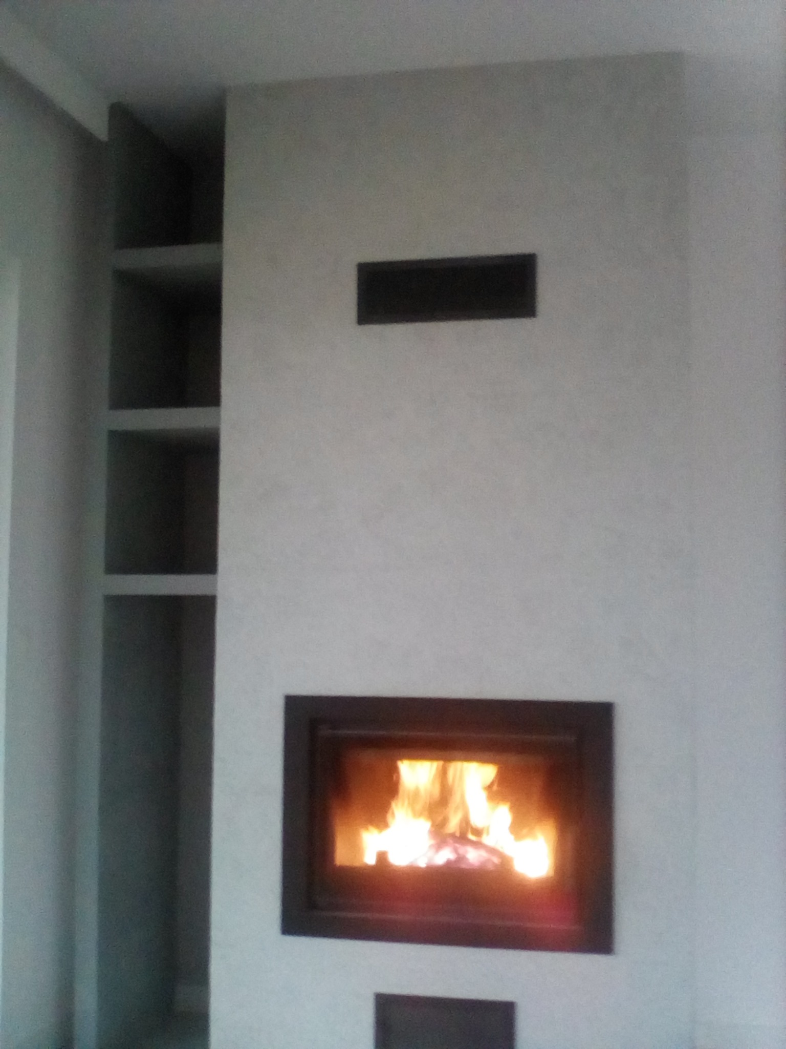 Wnętrze z kominkiem w zabudowie z szarej ściany z półkami po lewej stronie, widoczne płomienie.