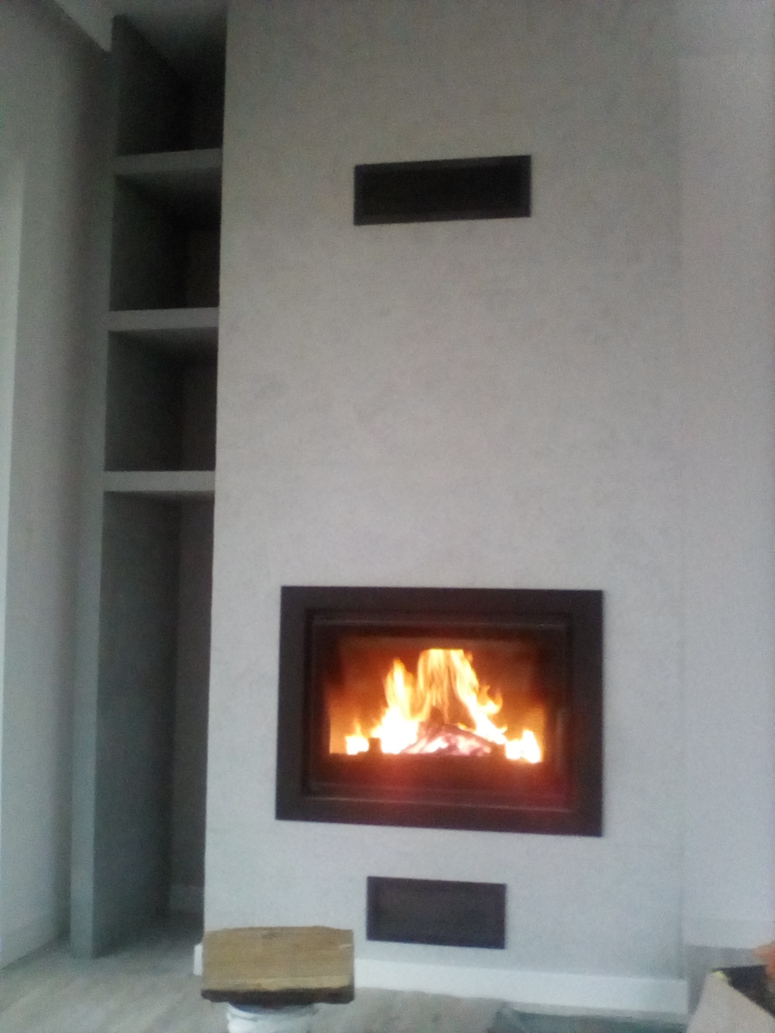 Minimalistyczny kominek z czarną ramą w ścianie pokrytej gładzią, płomienie widoczne przez szybę, obok wnęka z półkami, na podłodze widoczne ślady po remoncie.