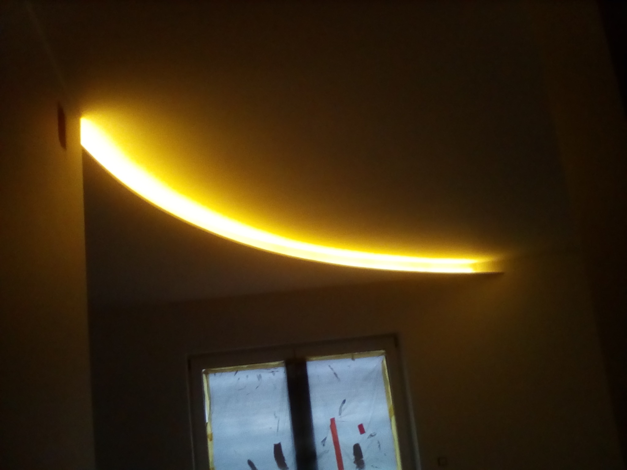 Zakrzywiona listwa LED, dająca ciepłe żółte światło, zamontowana na suficie w rogu pokoju, widoczna oklejona folią okienna rama.