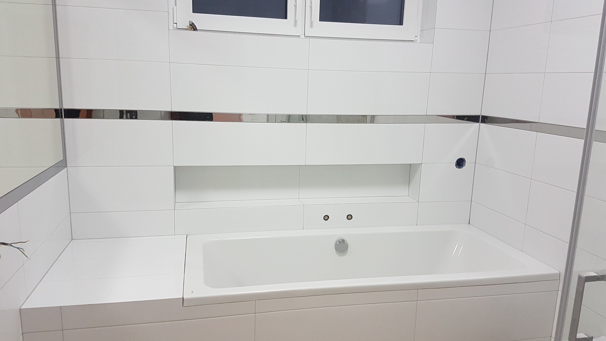Biała łazienka z wanną obudowaną płytkami, chromowane listwy dekoracyjne, okno w górnej części ściany, widoczne przyłącza hydrauliczne.