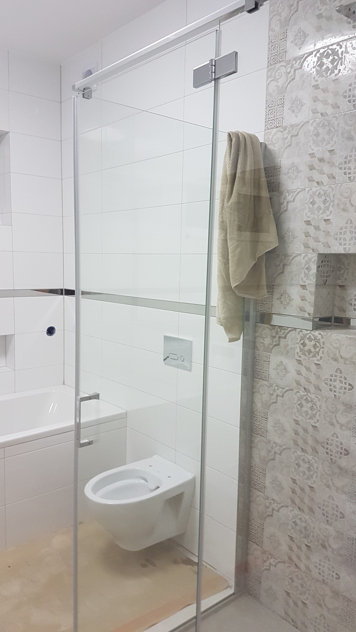 Nowoczesna łazienka z kabiną prysznicową ze szkła hartowanego, wisząca toaleta, białe i wzorzyste płytki oraz beżowy ręcznik na relingu.