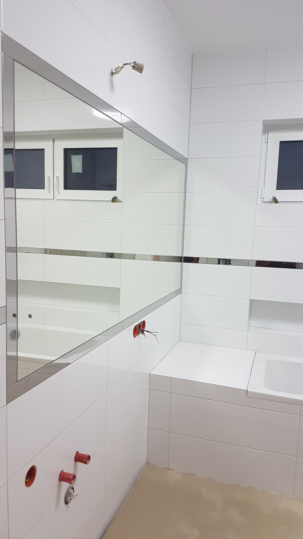 Biała łazienka w trakcie wykańczania, widoczne białe płytki, lustro z metalową ramą, przygotowane punkty instalacji elektrycznej i wodnej, zabezpieczona podłoga.