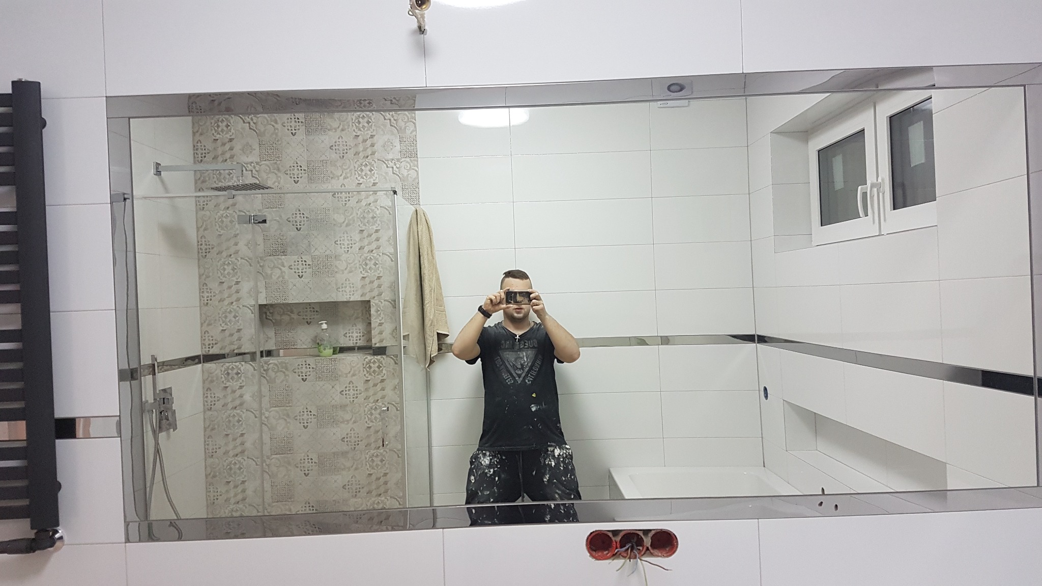 Odbicie w lustrze łazienki po remoncie, widoczna kabina prysznicowa z wzorzystymi płytkami, białe płytki ścienne z metalowymi listwami, okno, oraz osoba robiąca zdjęcie telefonem, ubrana w robocze...