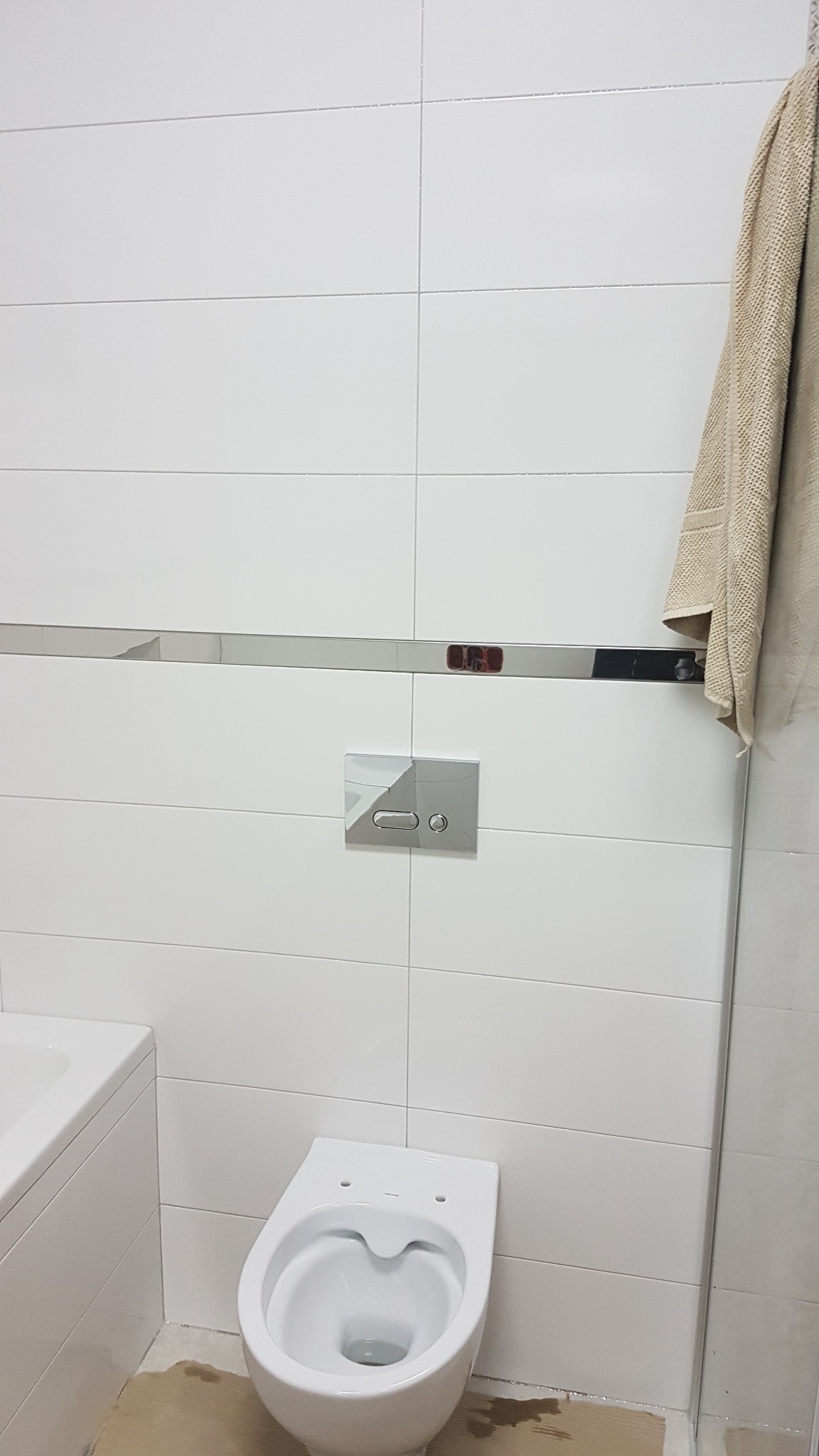 Biała wisząca toaleta bez deski, zamontowana na ścianie z białych płytek z chromowaną listwą dekoracyjną i przyciskiem spłukującym, obok fragment wanny i beżowy ręcznik.