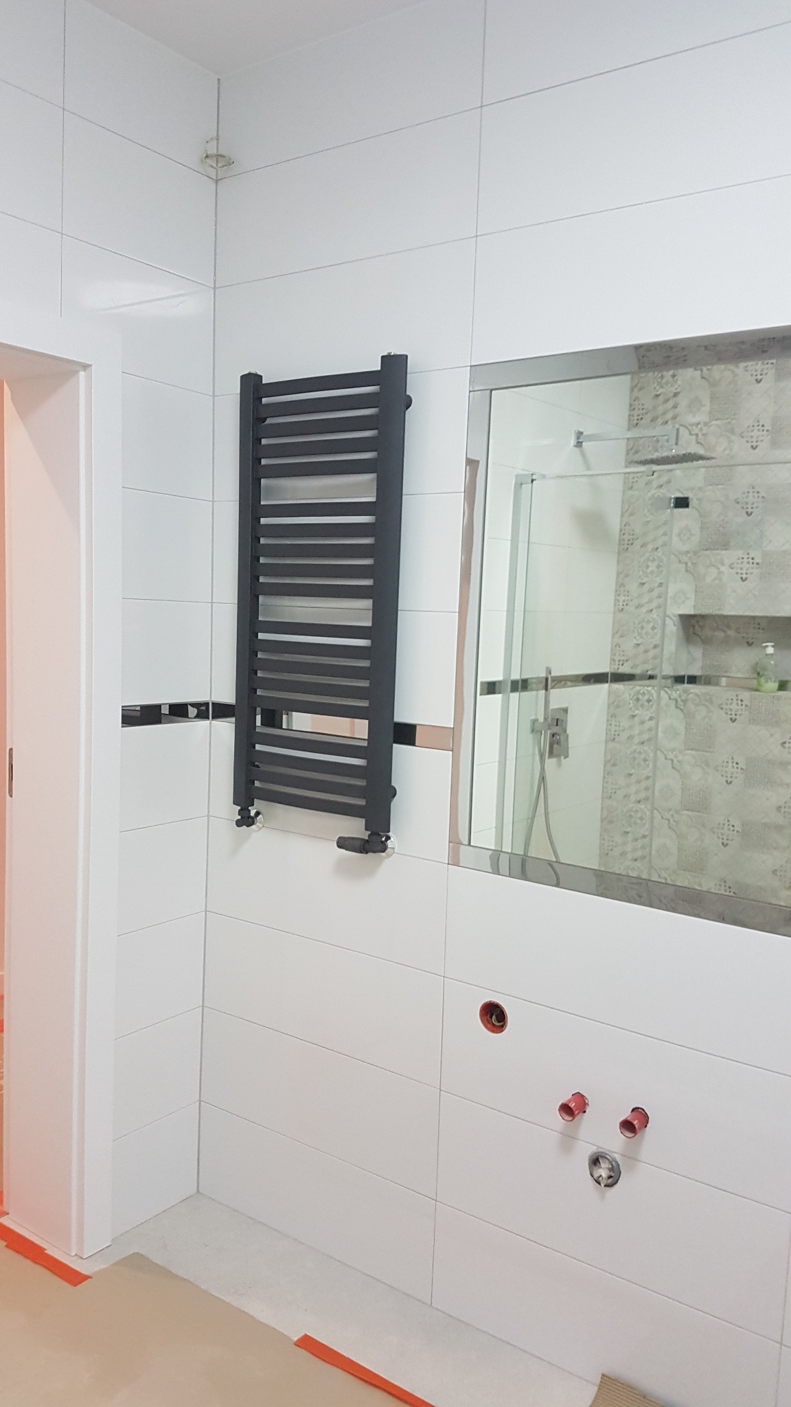 Łazienka w trakcie wykończenia: białe płytki, zamontowany grafitowy grzejnik drabinkowy, lustro z widocznym prysznicem w odbiciu, przygotowane podejścia pod armaturę, zabezpieczona podłoga.