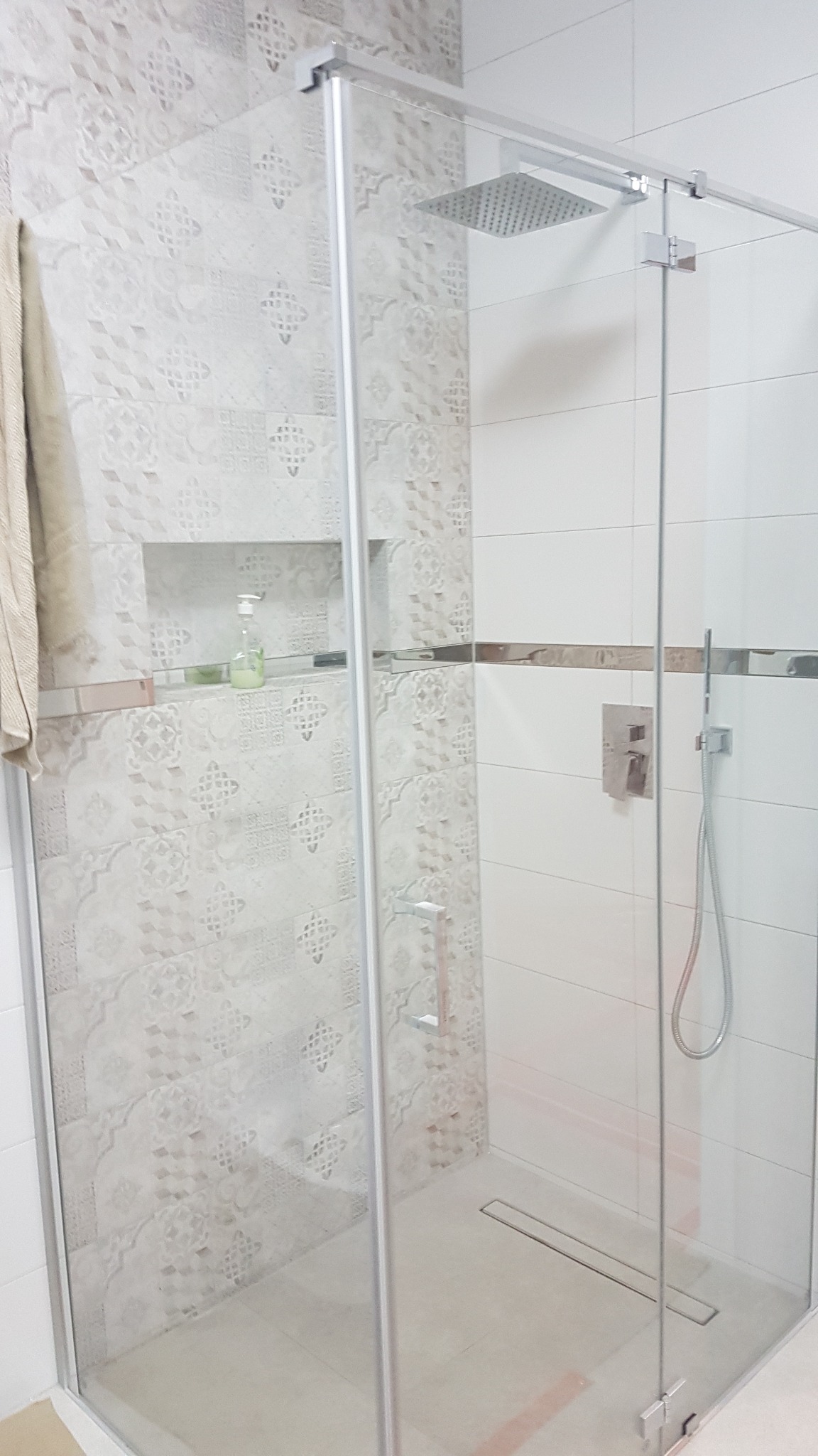 Nowoczesna kabina prysznicowa z deszczownicą i szklanymi drzwiami, wykończona płytkami w odcieniach bieli i beżu z dekoracyjnym wzorem. W ścianie wnęka z kosmetykami.