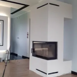 Nowoczesny kominek obudowany płytą karton gips, pomalowany na biało, z czarnym wkładem kominkowym, widoczny w salonie z drewnianą podłogą i dekoracyjnym oświetleniem sufitowym.