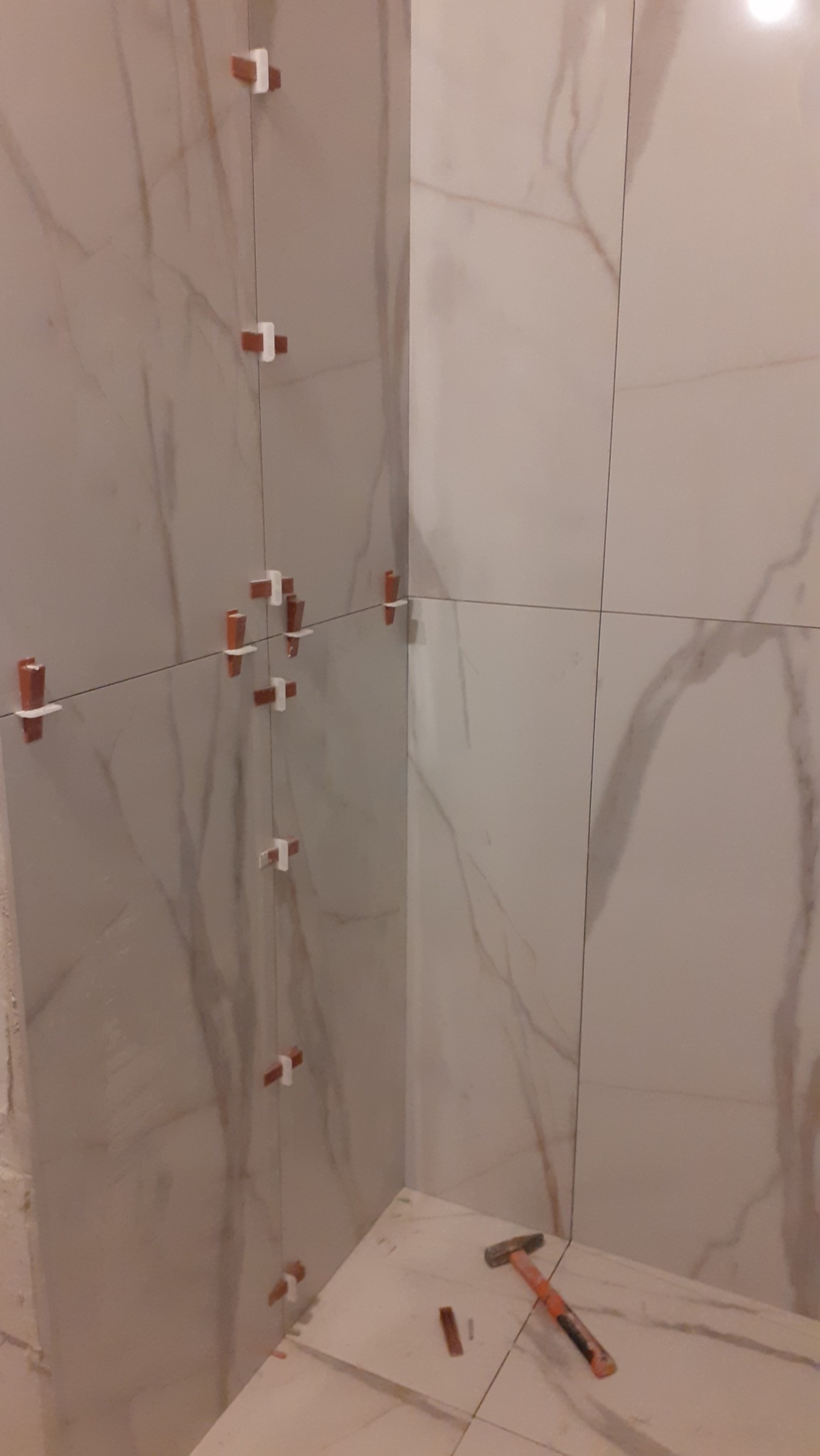 Ściana w trakcie układania płytek imitujących marmur, z widocznymi krzyżykami dystansowymi i gumowym młotkiem na podłodze. Częściowo ułożona podłoga.