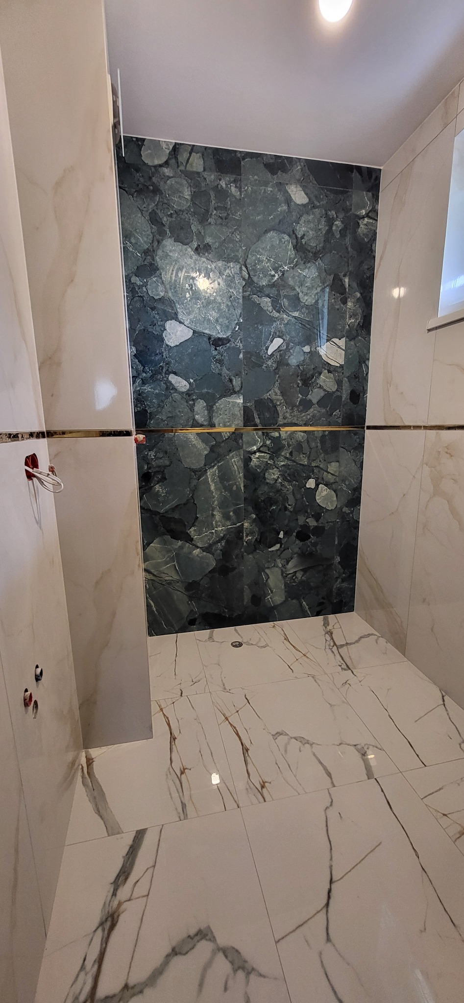 Wykończona łazienka z marmurowymi płytkami w odcieniach bieli i zieleni. Widoczne przyłącza hydrauliczne i odpływ podłogowy.